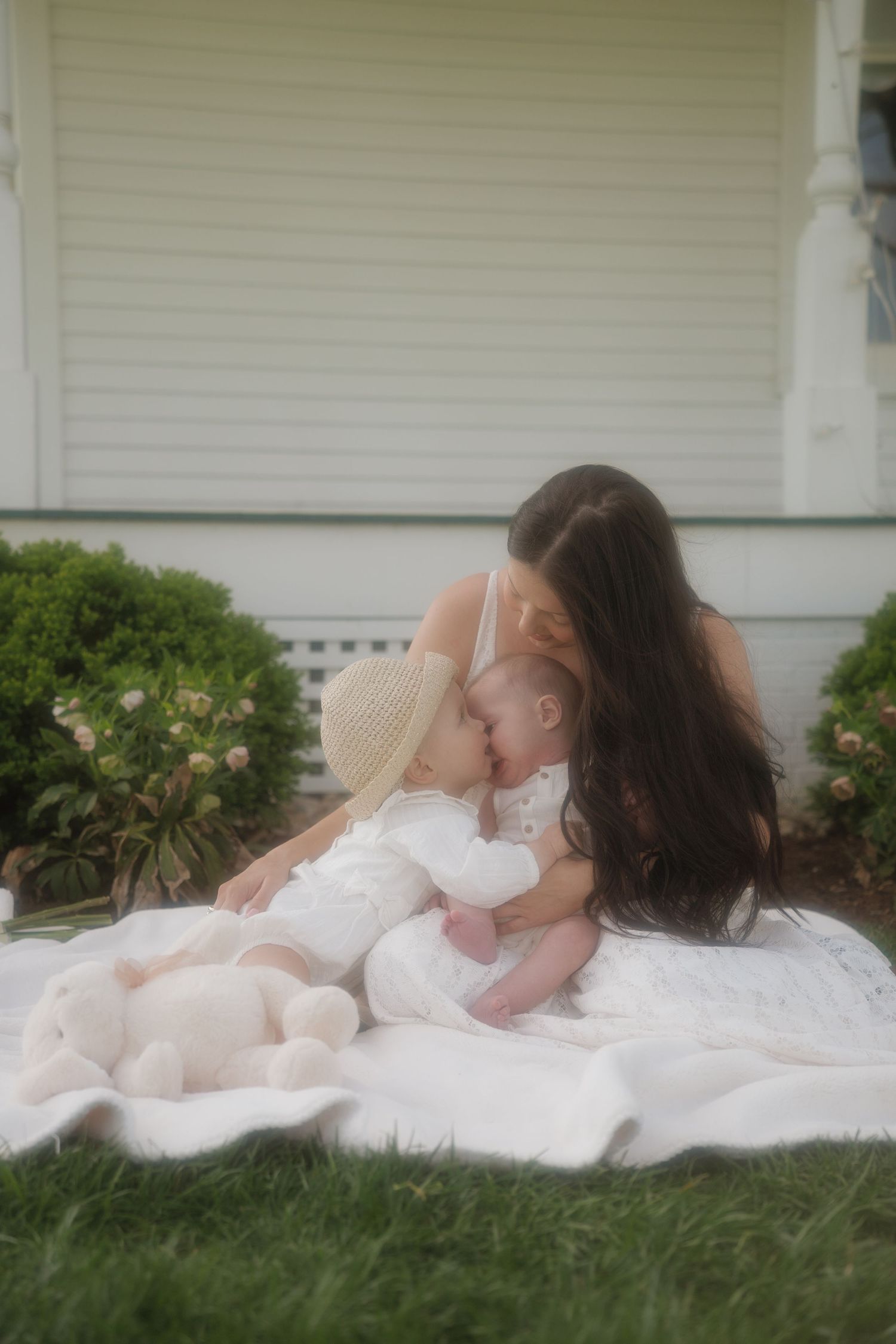 A mother in white dress kisses her baby on a white blanket in front of a house.