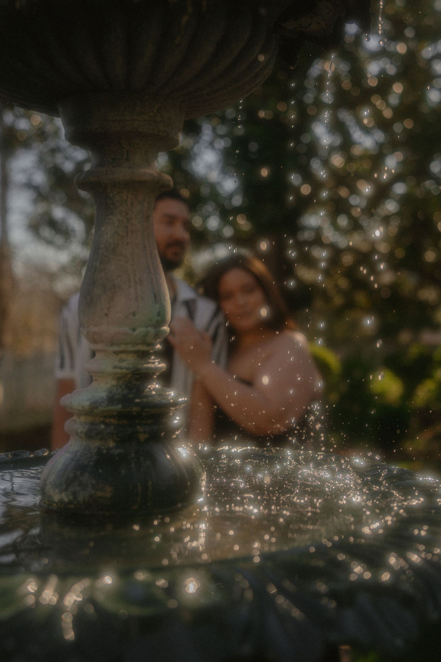 Romantic couple blurred behind a stone garden fountain with sparkling water droplets.