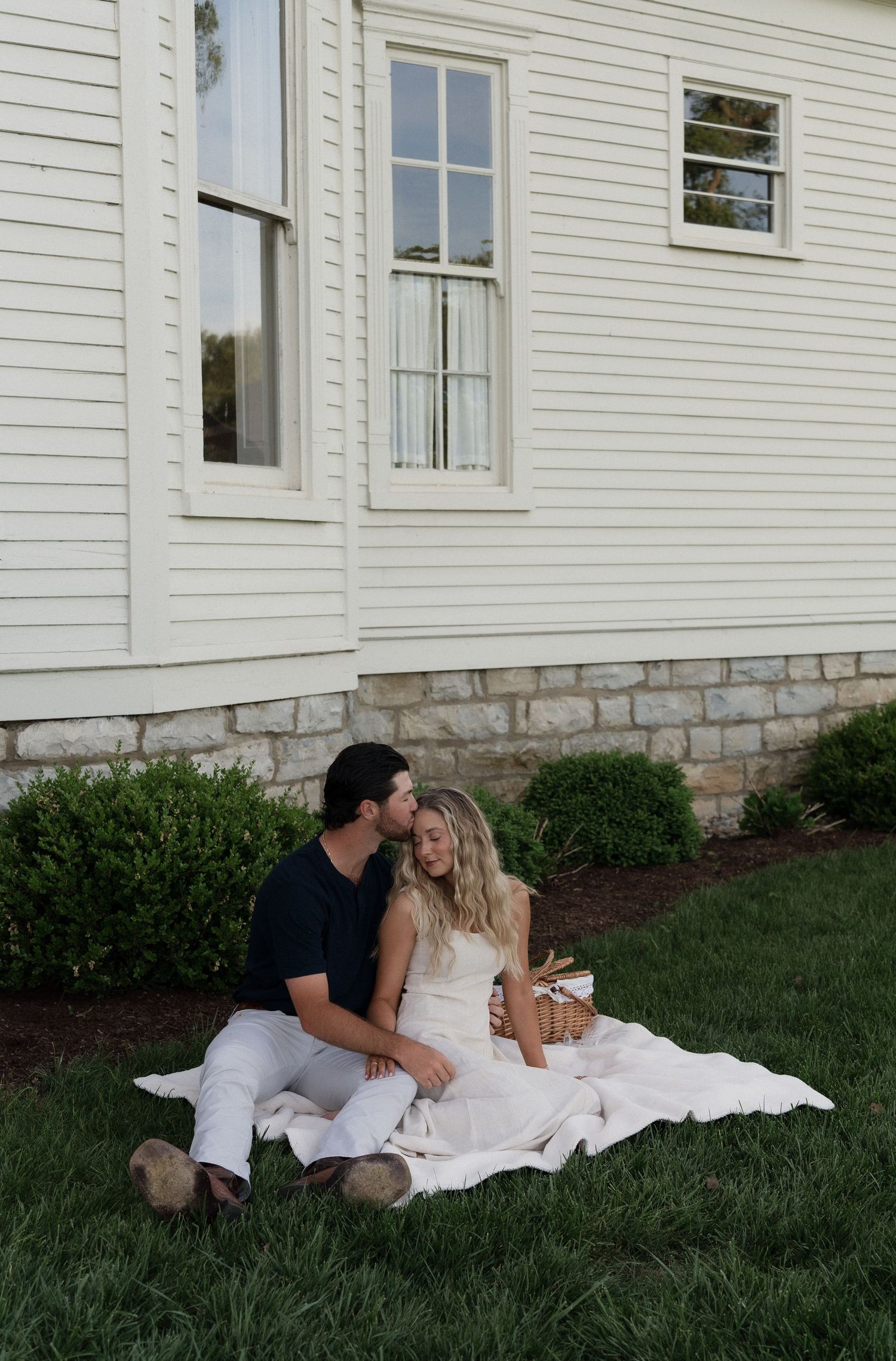 Couple sitting on white blanket in grass beside white farmhouse, sharing a tender kiss.