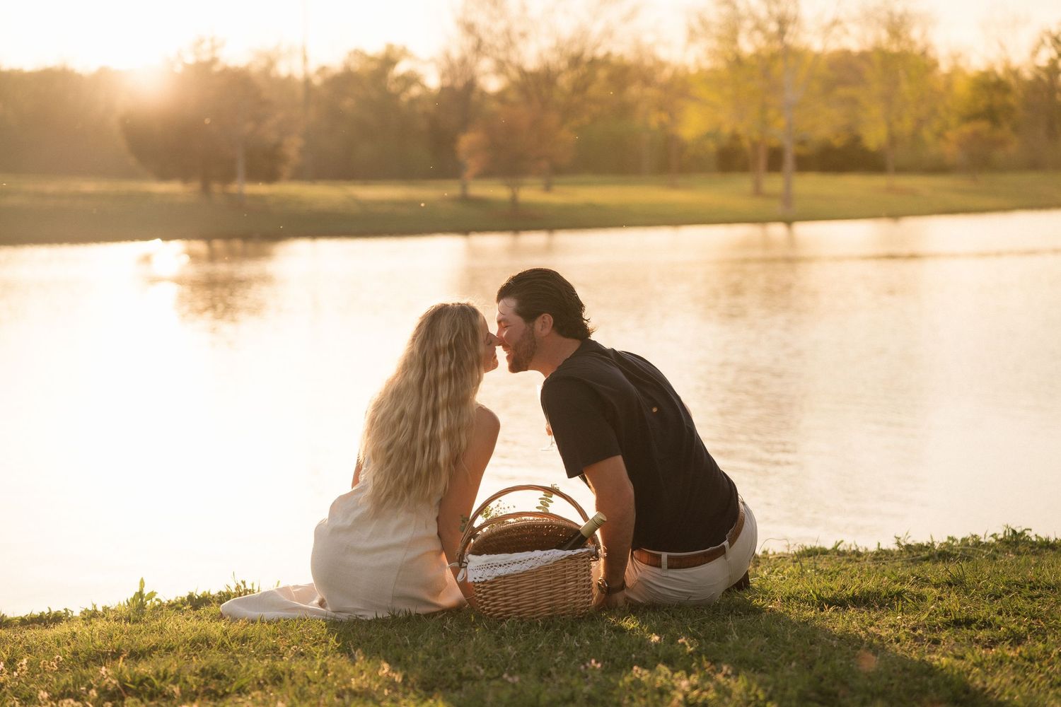 Couple sharing a romantic kiss during a golden sunset picnic by a serene lake with a wicker basket.