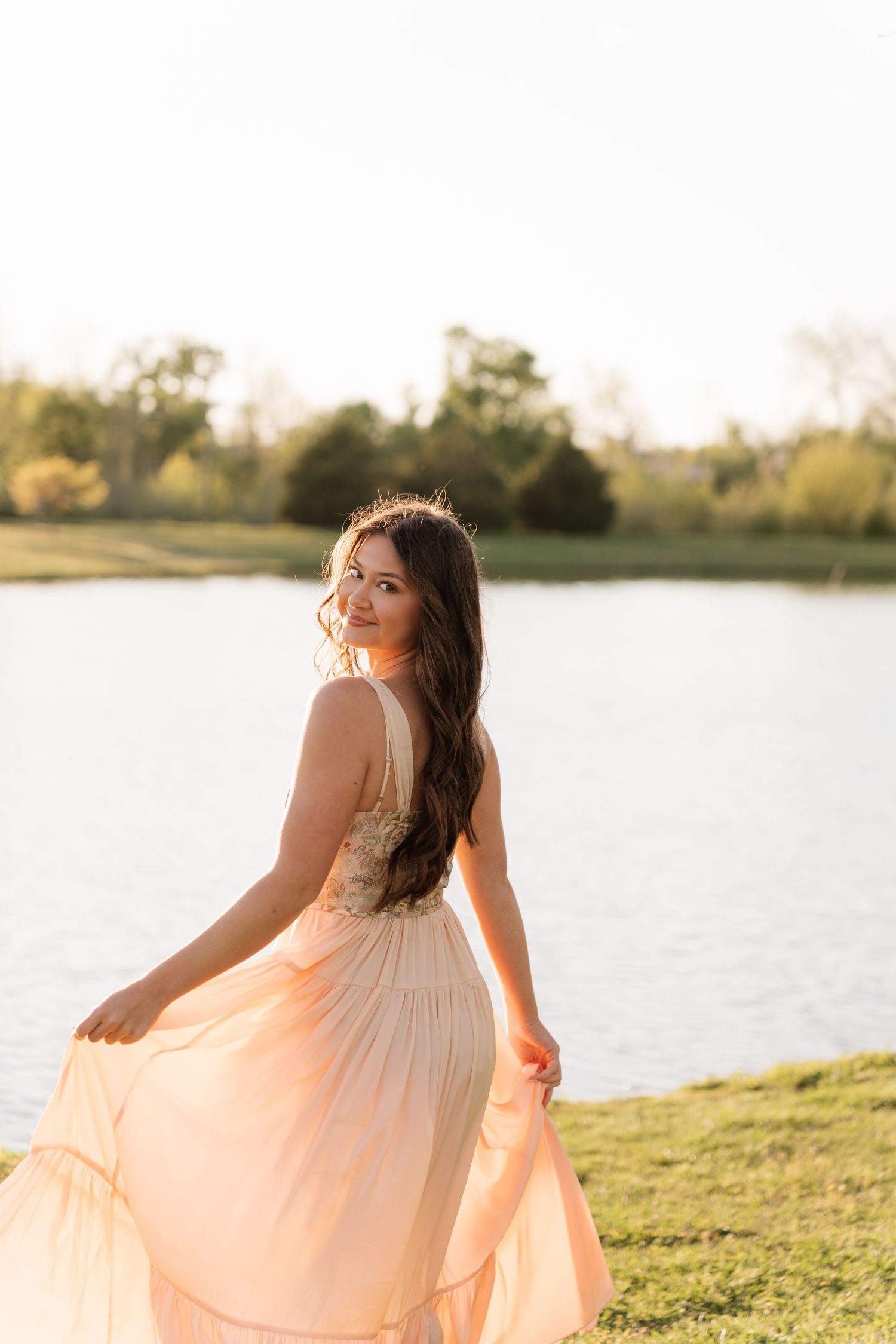 Brunette woman in flowing peach dress twirling by lakeside in warm golden sunset light.