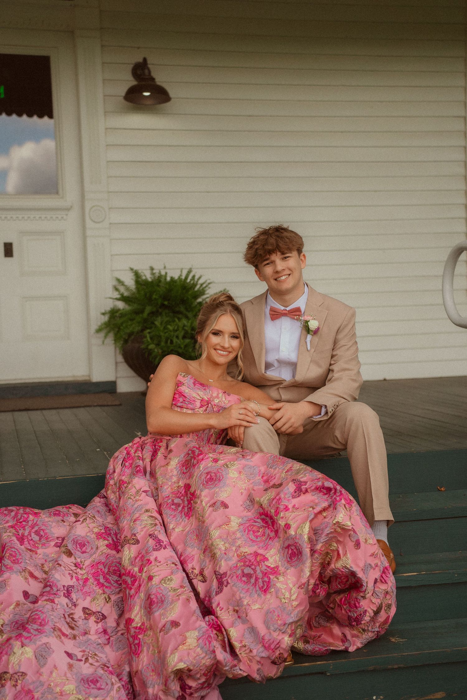 A young couple poses on porch steps, girl in pink floral gown, boy in tan suit with pink bow tie.