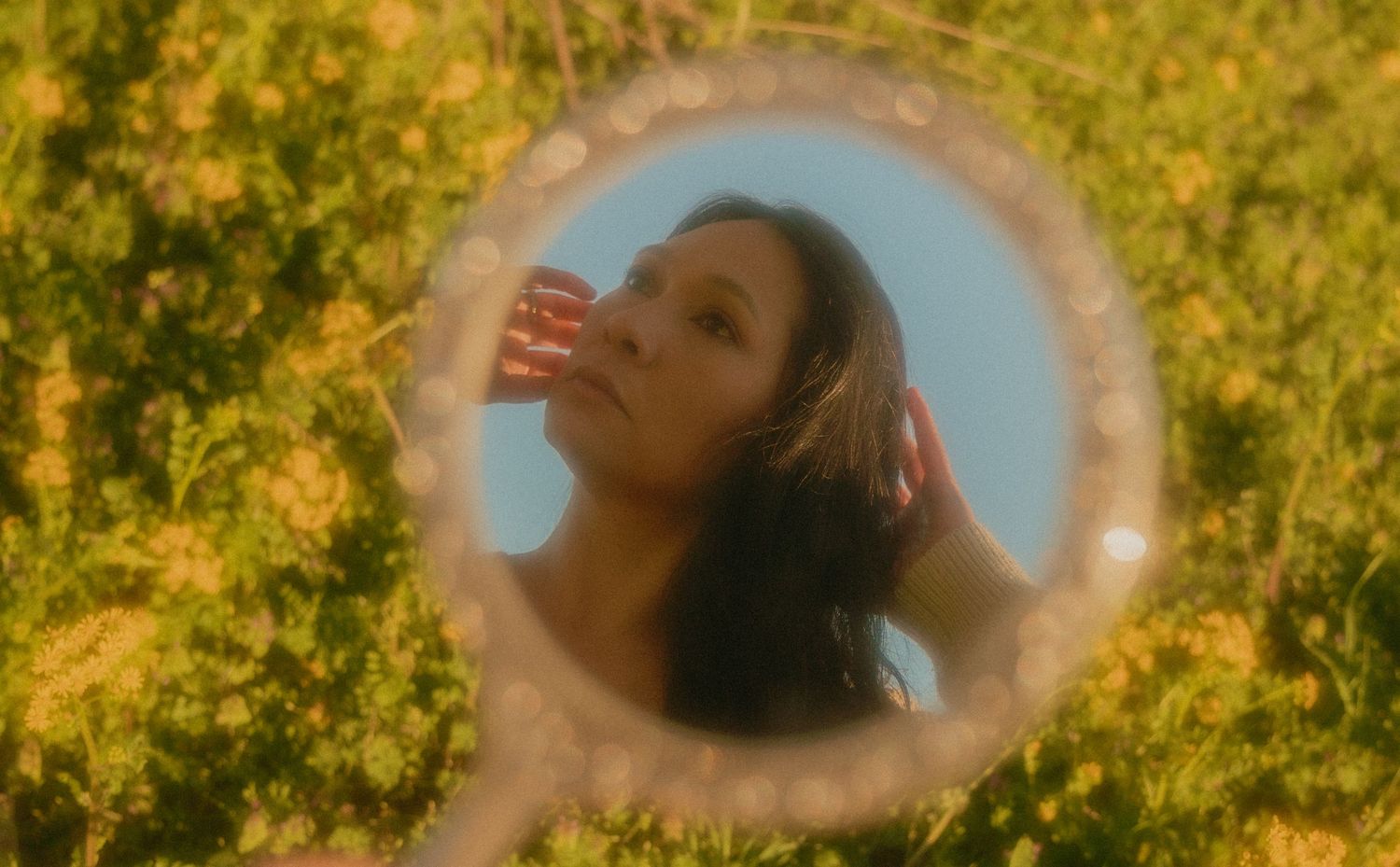 Woman reflected in oval mirror surrounded by yellow wildflowers against a blue sky.