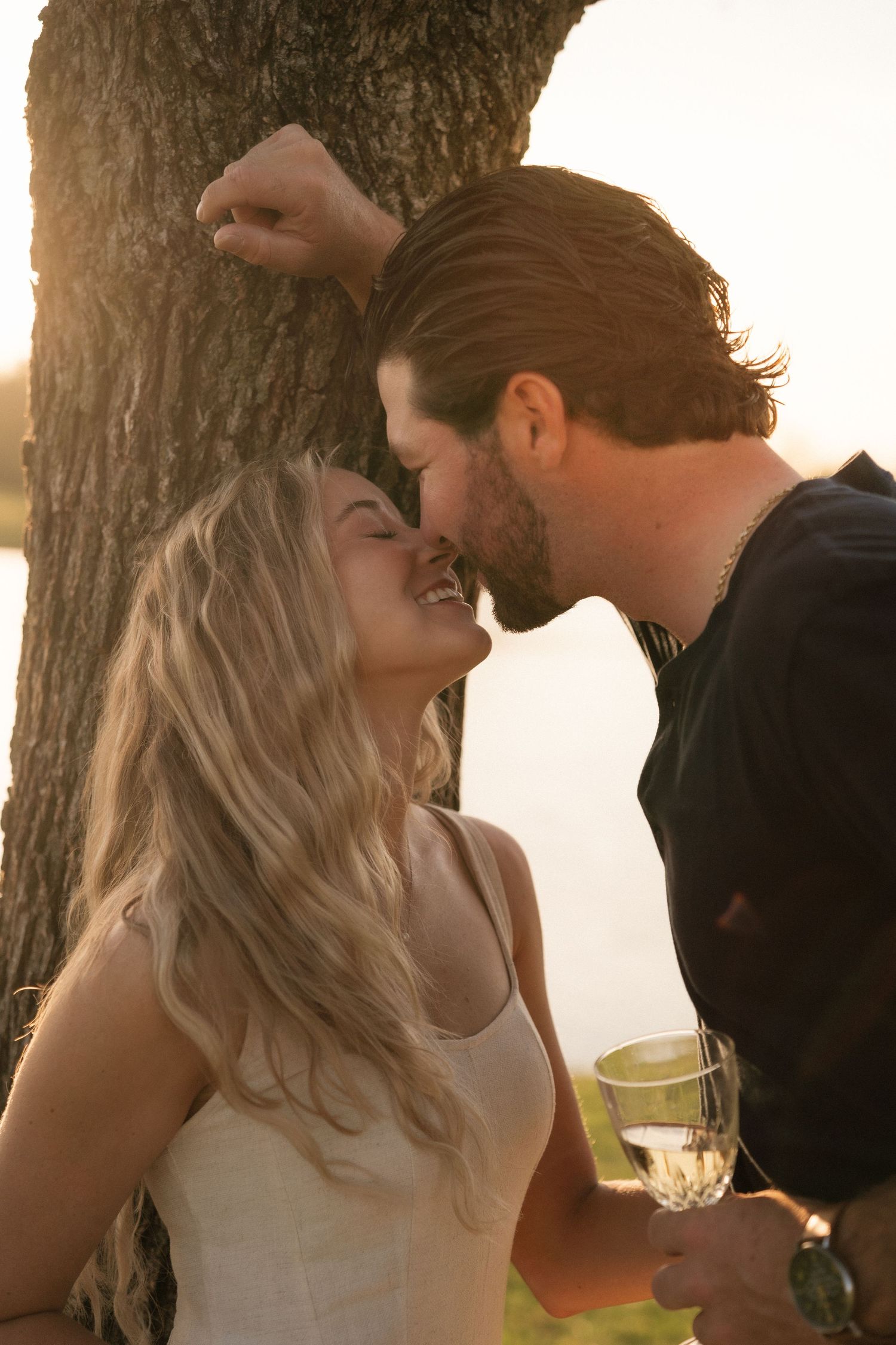 A romantic couple shares an intimate moment near a tree at sunset, with the man holding a glass of wine.