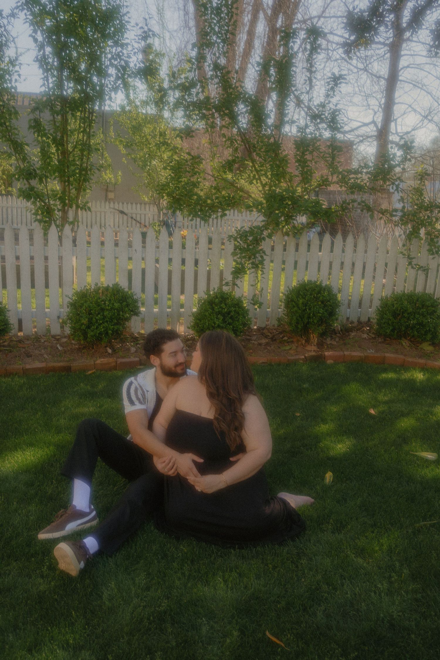 Couple sitting on green grass in a backyard, sharing a romantic moment near a white picket fence and lush shrubs.