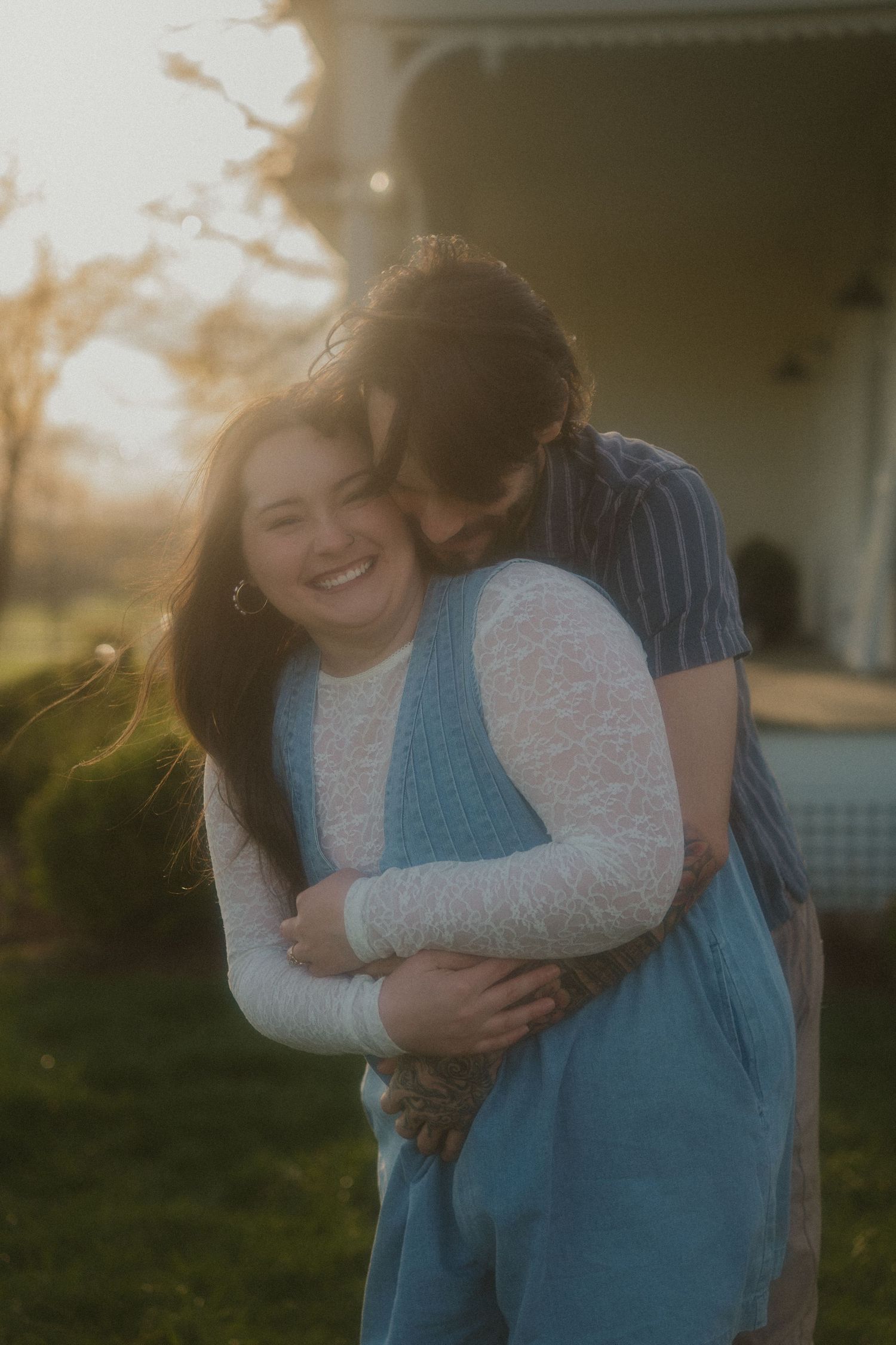 Couple embracing at golden hour outside a farmhouse, woman smiling joyfully as man hugs her from behind.