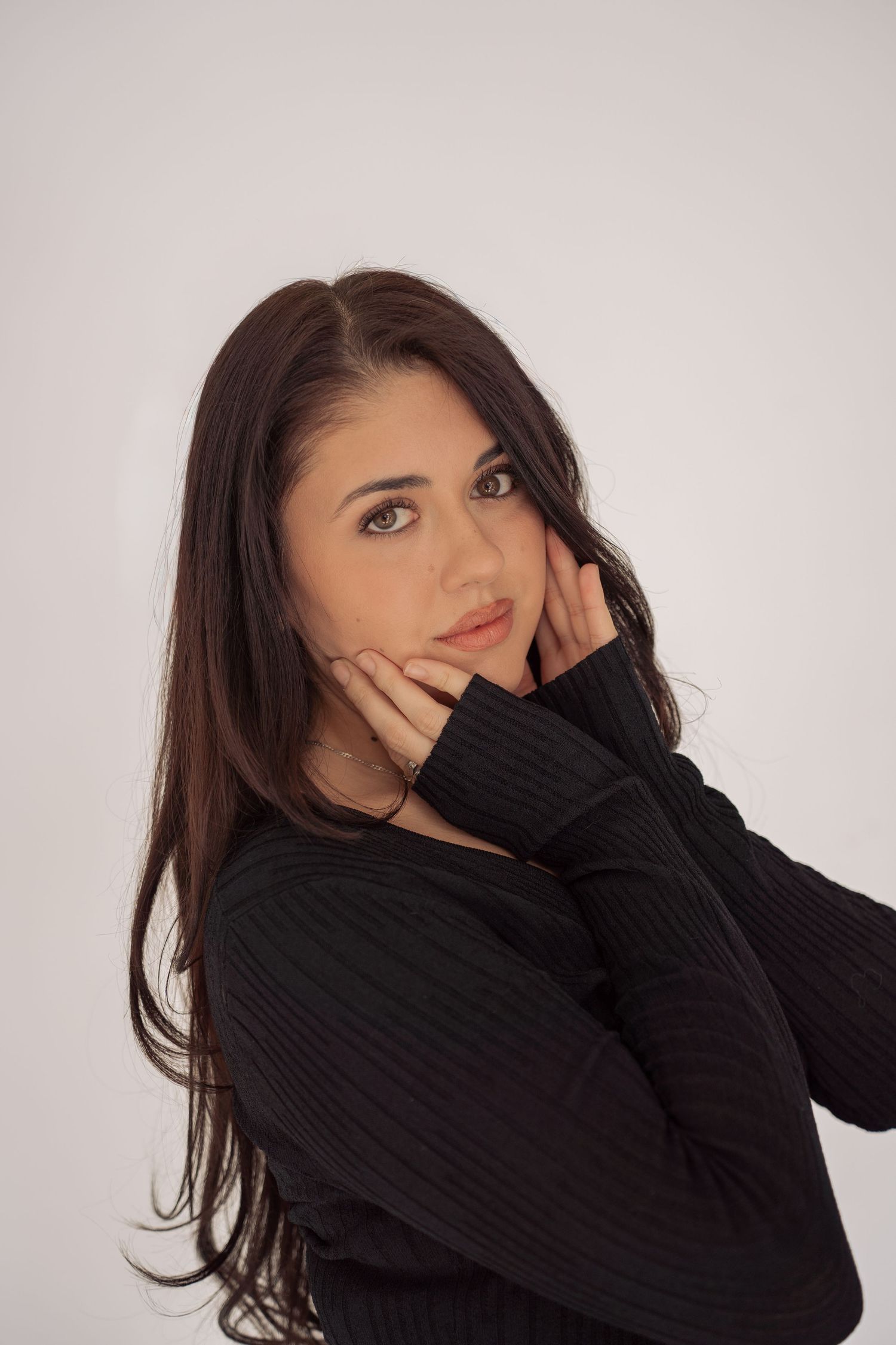 Young brunette woman in black ribbed sweater posing with hands near face against white background.