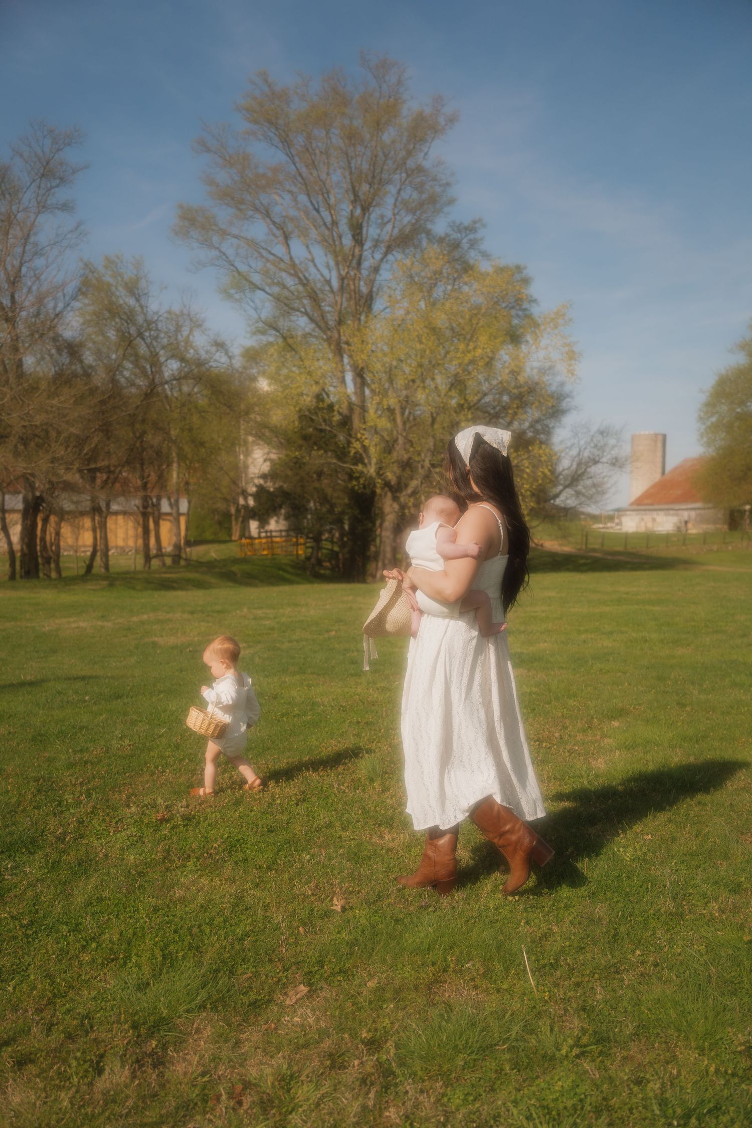 Mother in white dress and boots holding newborn while toddler walks nearby in sunny green field.