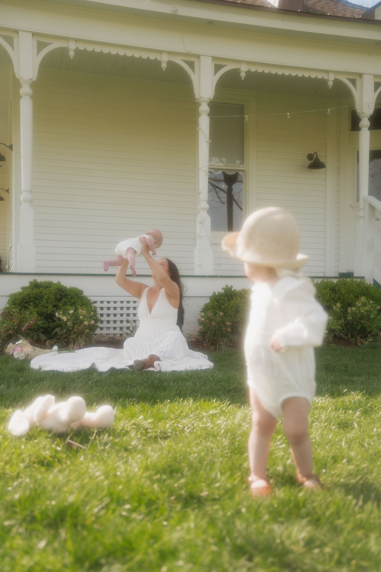 Mother lifting baby in air on lawn in front of white Victorian farmhouse with toddler in hat nearby.
