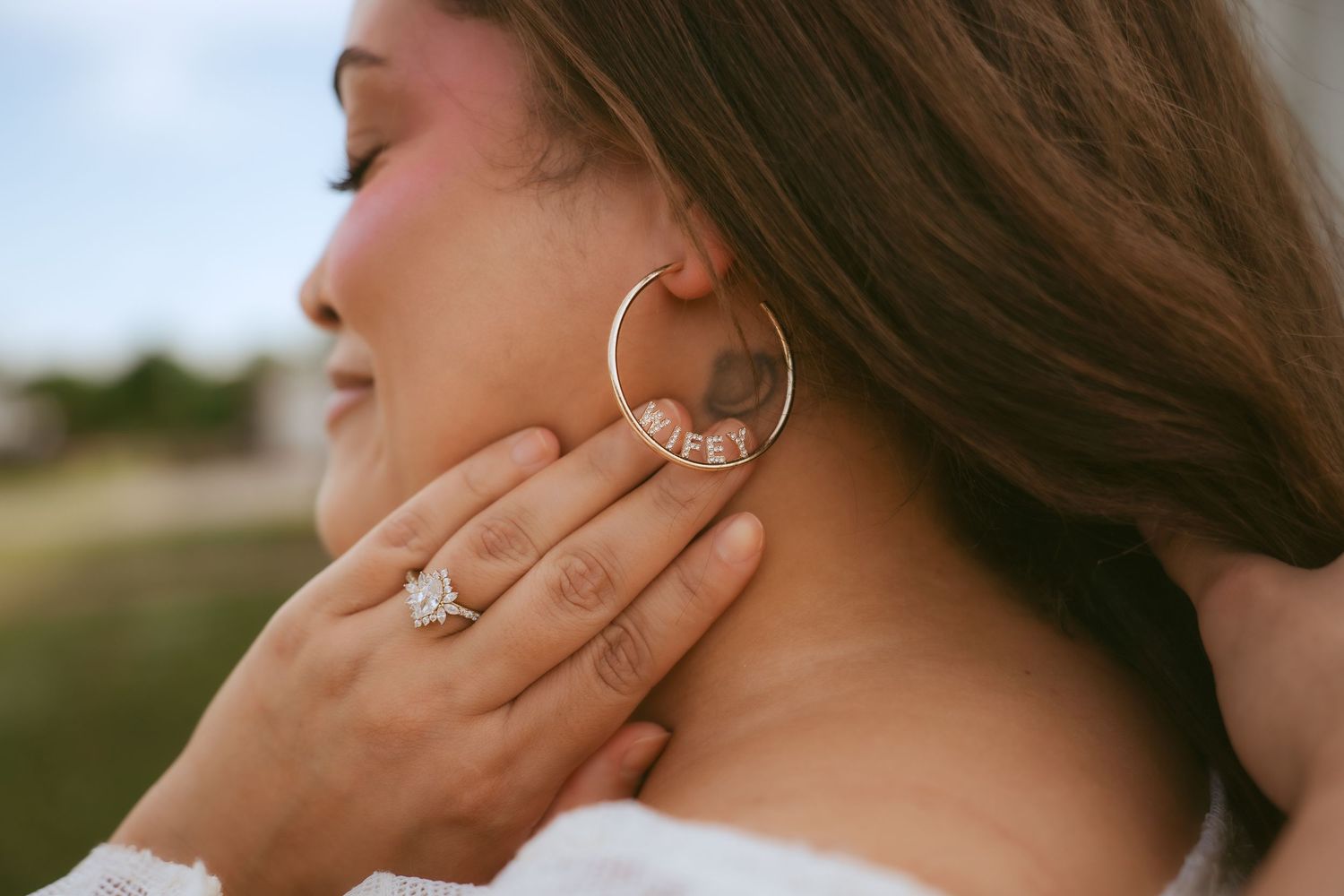 Close-up of a woman wearing a gold hoop earring reading WIFEY and a diamond engagement ring.