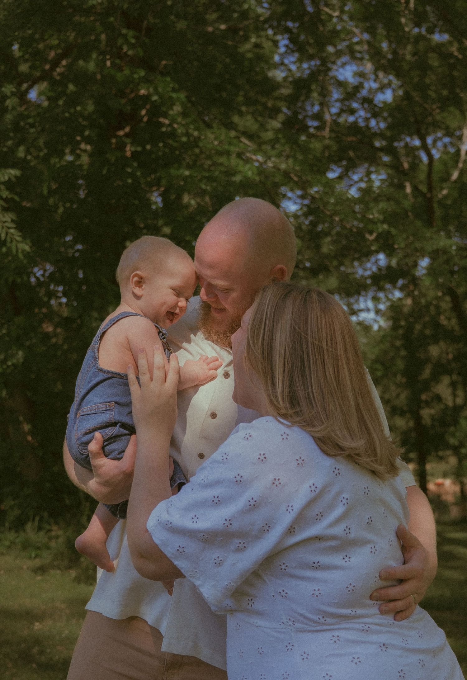 A bald man and blonde woman share a joyful outdoor moment with their laughing baby in a sunny green park.