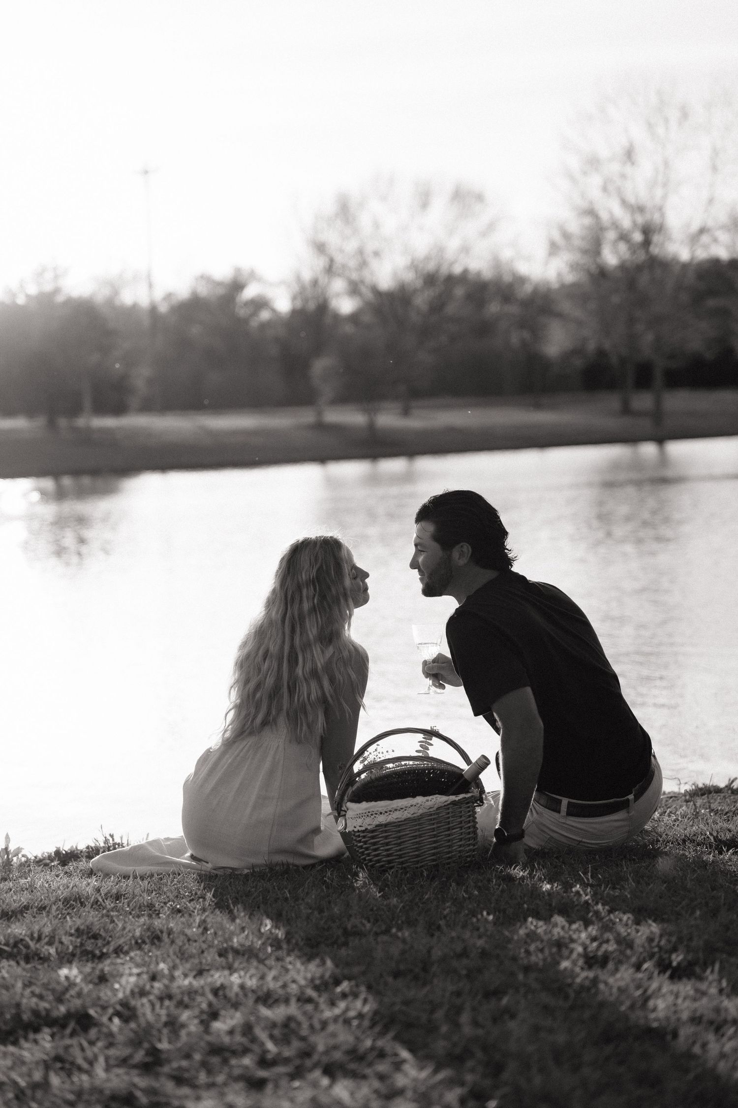 Black and white photo of couple sitting together by lake with picnic basket in golden light.