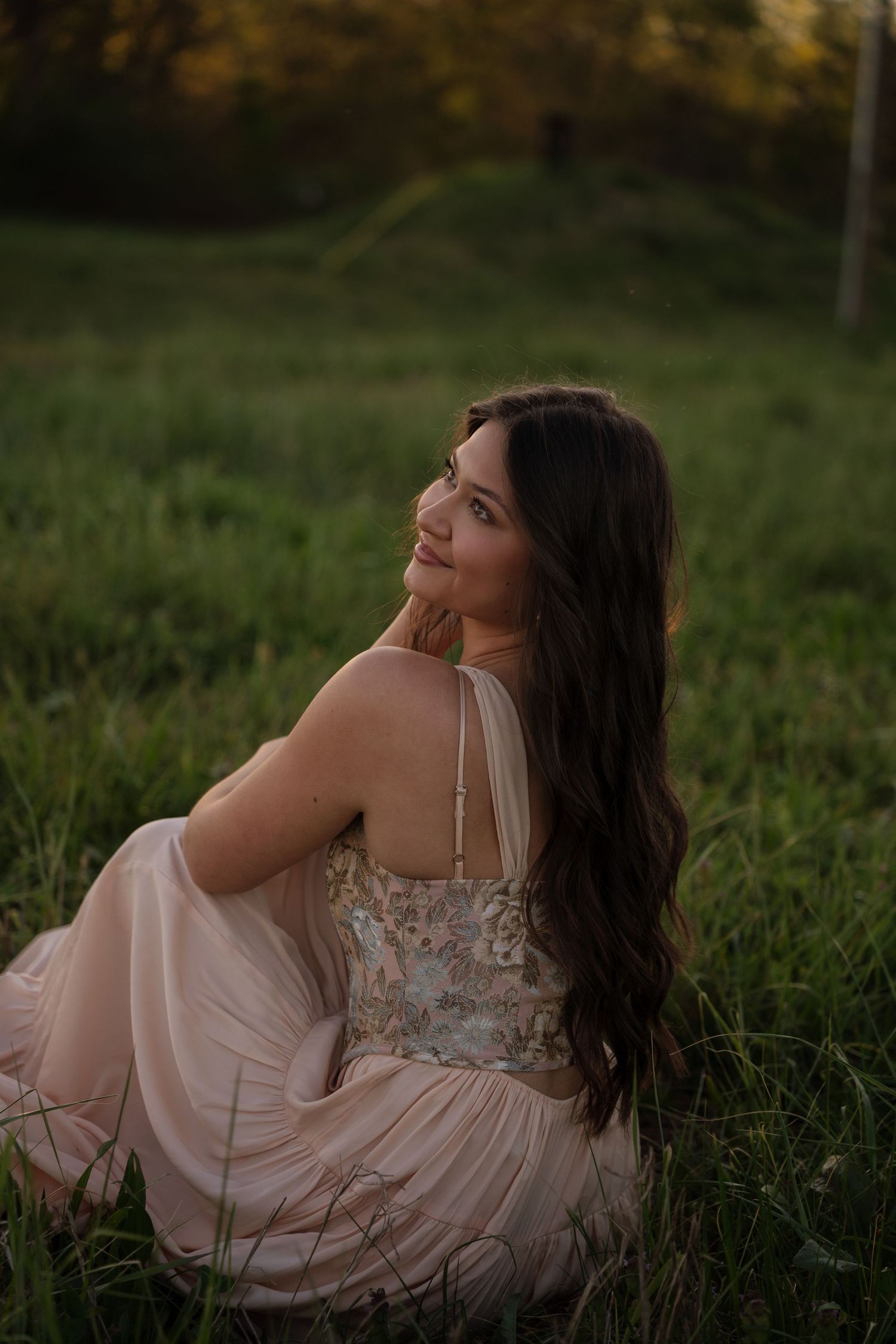 Young woman in floral pink dress sitting in green grass field at golden hour, smiling over shoulder.