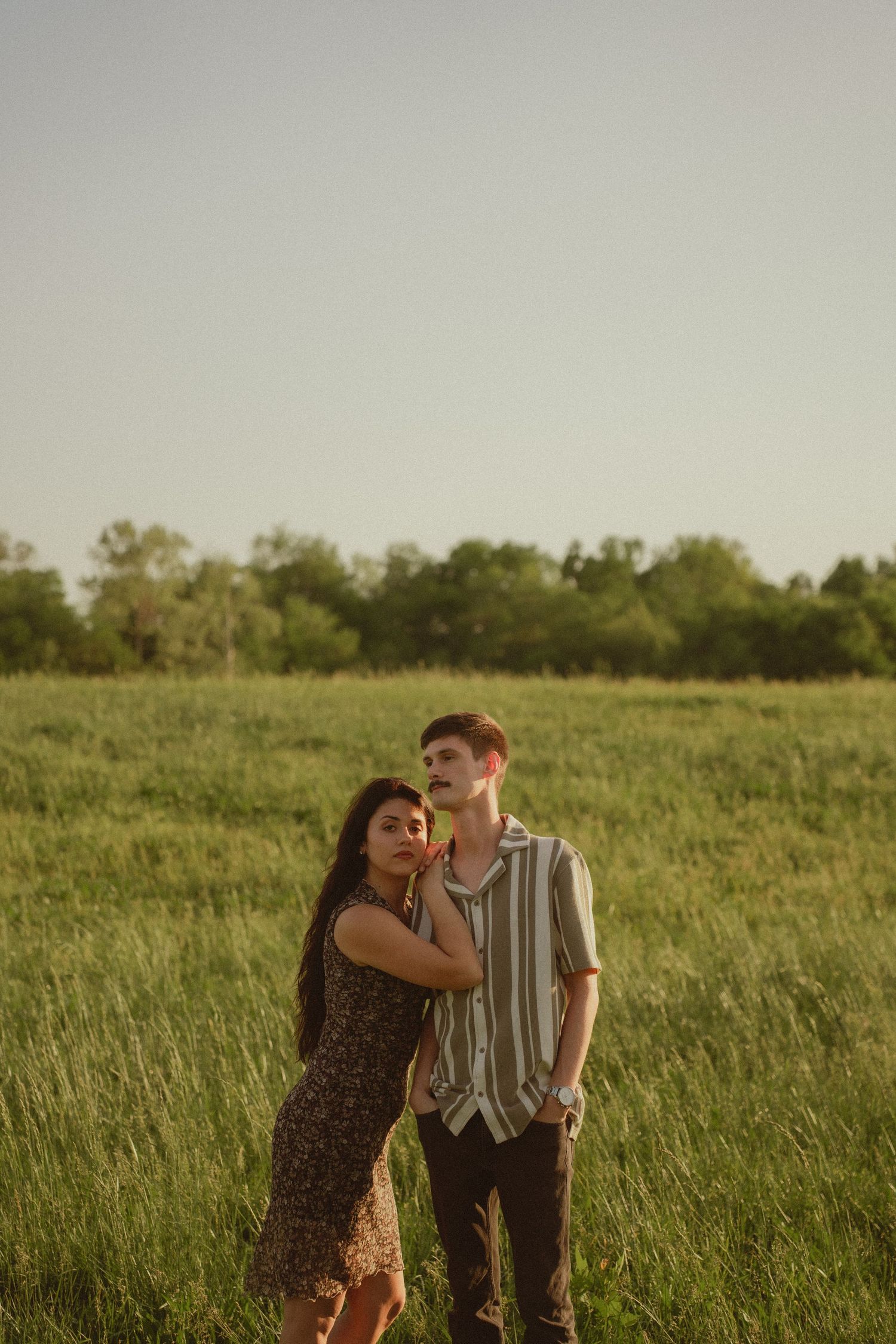 A young couple stands in a lush green field, embracing during golden hour with trees in the background.