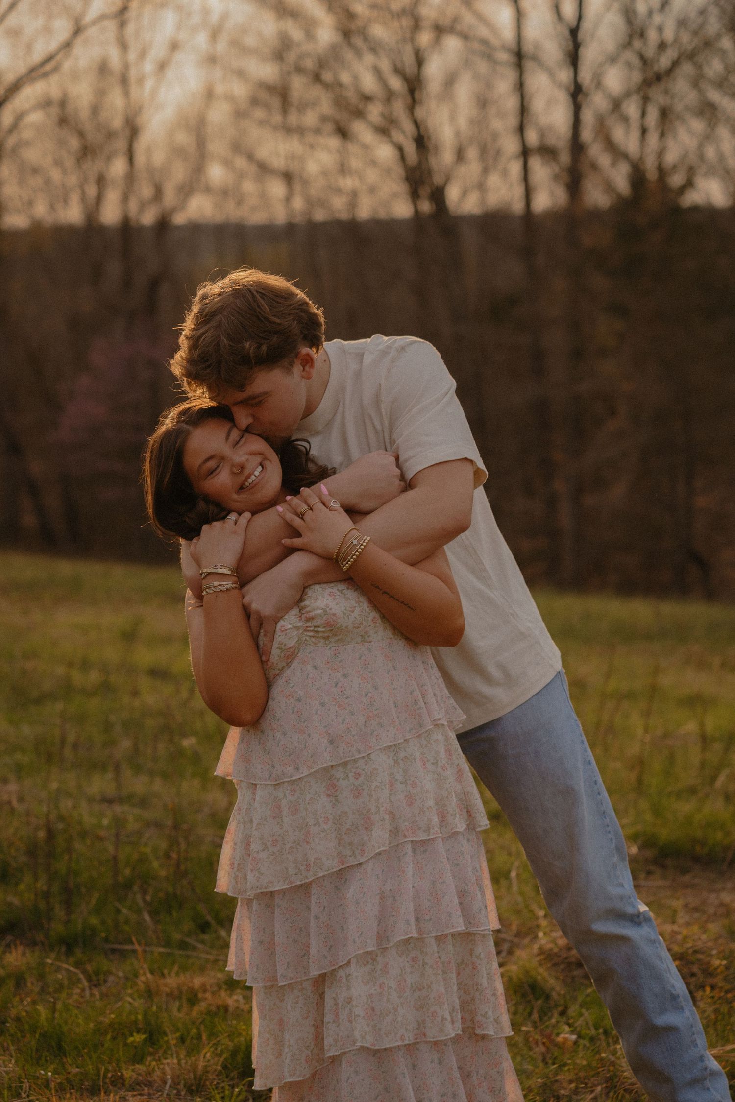 Couple embracing at golden hour sunset in a field, man hugging woman in floral tiered dress.