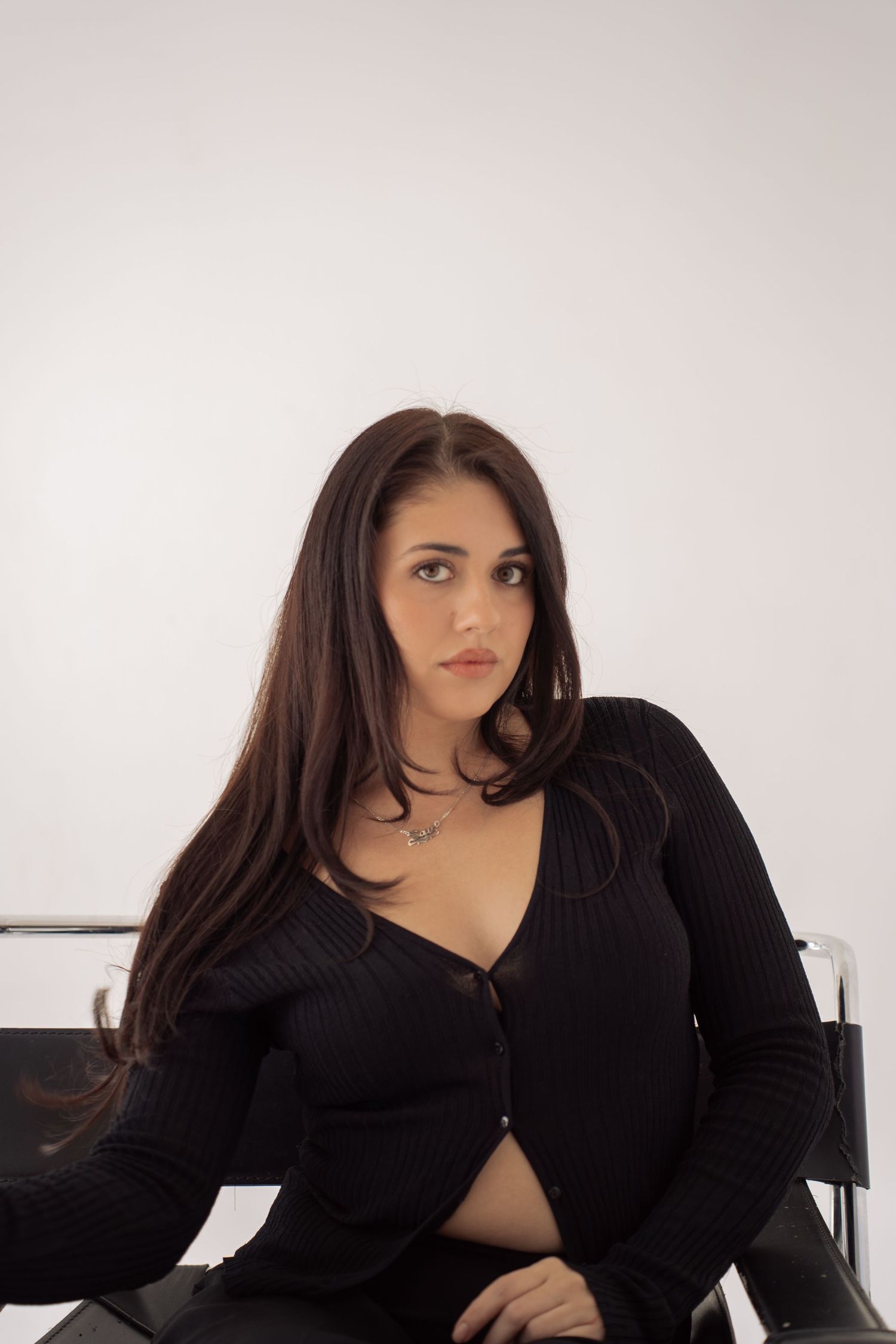 Young woman with long dark hair in black cardigan sitting in a modern chair against white background.