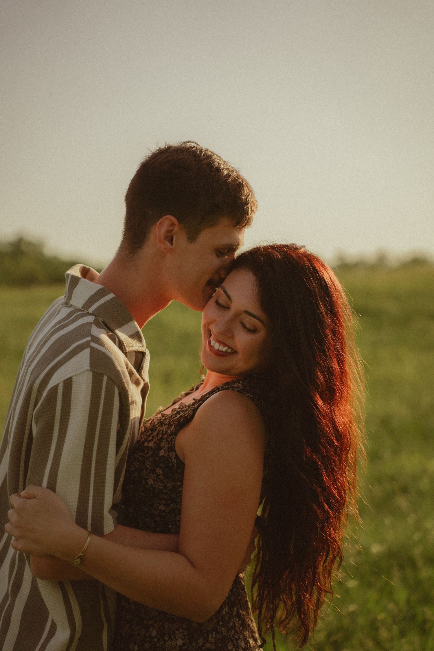 A couple embraces in a sunlit field, the woman smiling joyfully with red hair as the man leans in close.