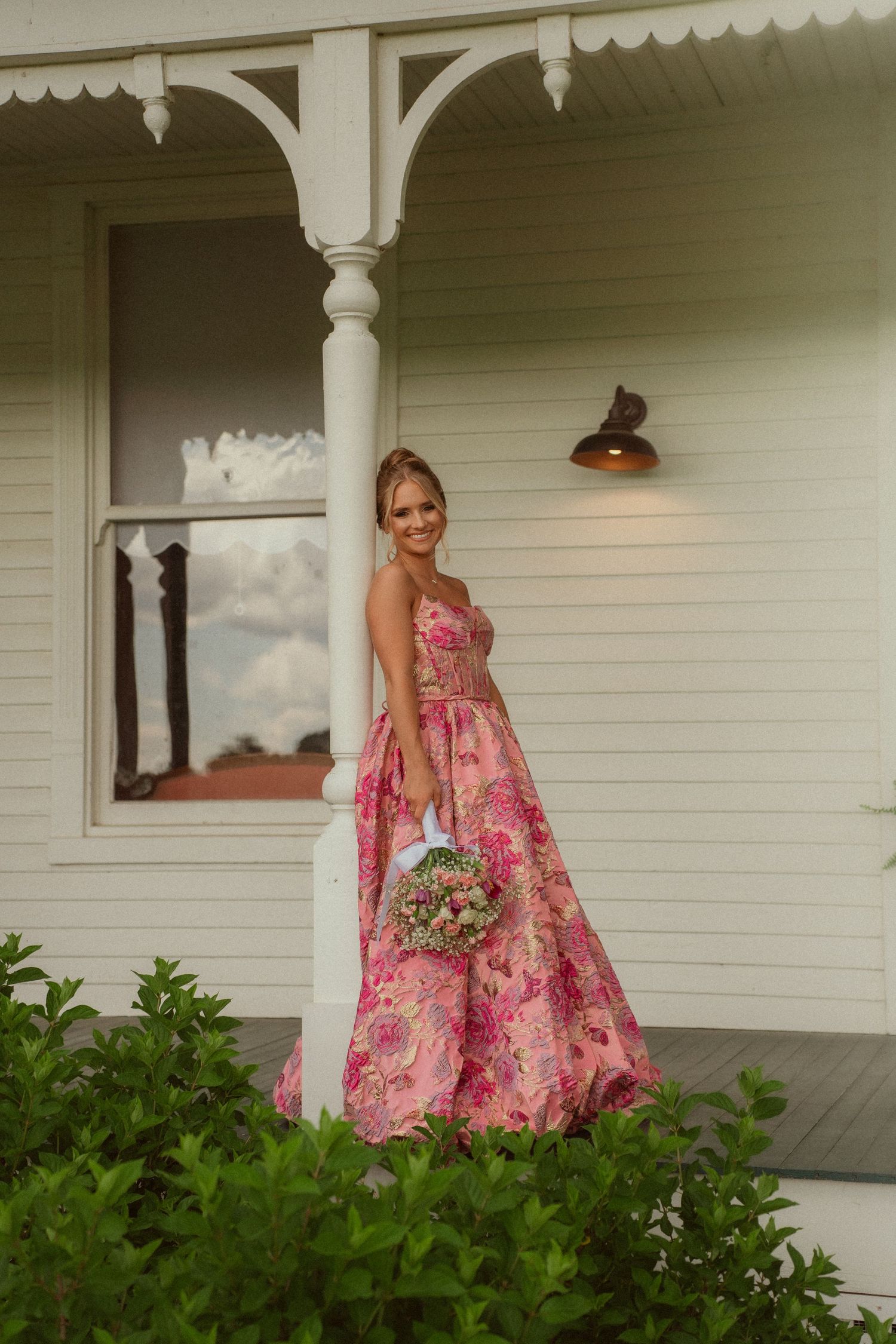 Woman in pink floral gown holding bouquet poses by white colonial porch column surrounded by lush greenery.