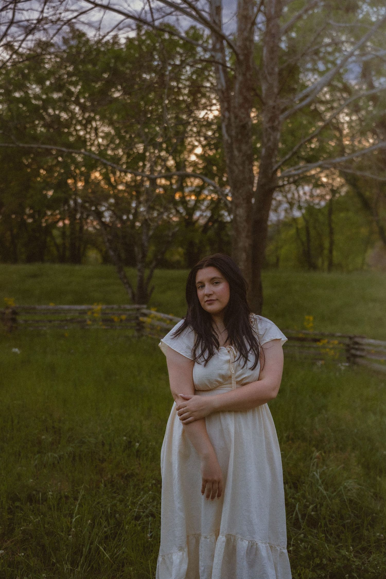 Young woman in white dress standing in green field at sunset with trees and wooden fence.