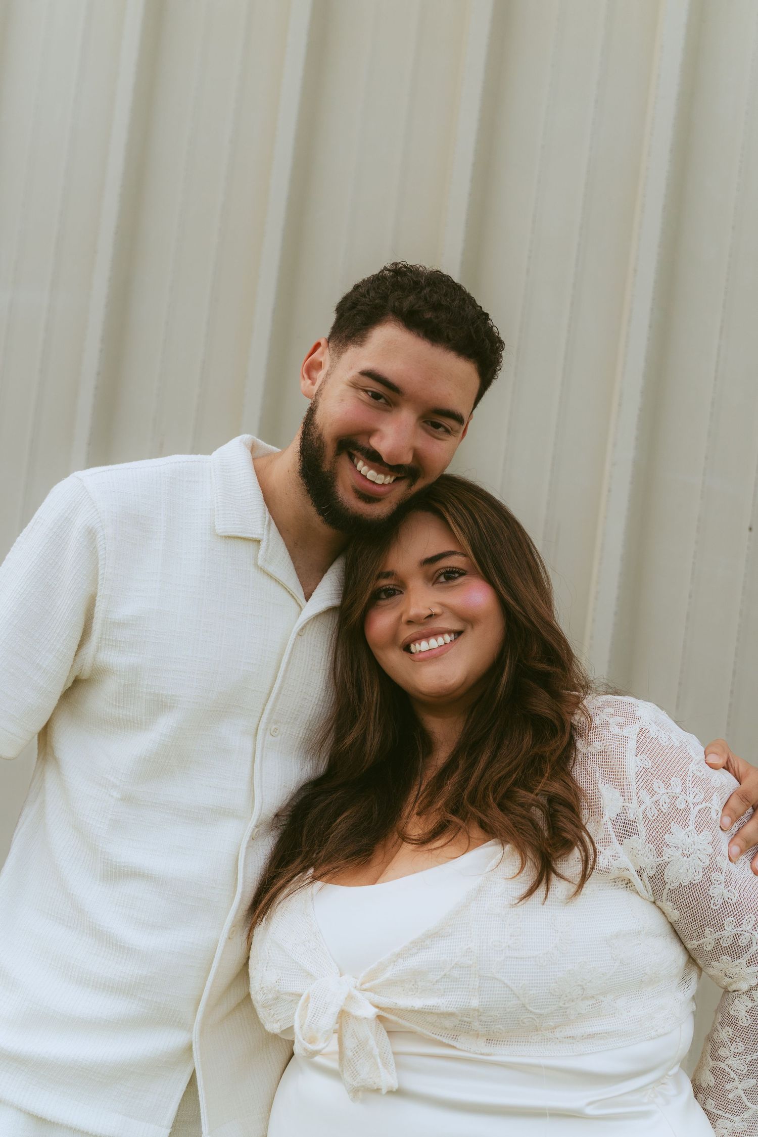 A smiling couple in matching white outfits poses together against a neutral background in a romantic portrait photo.