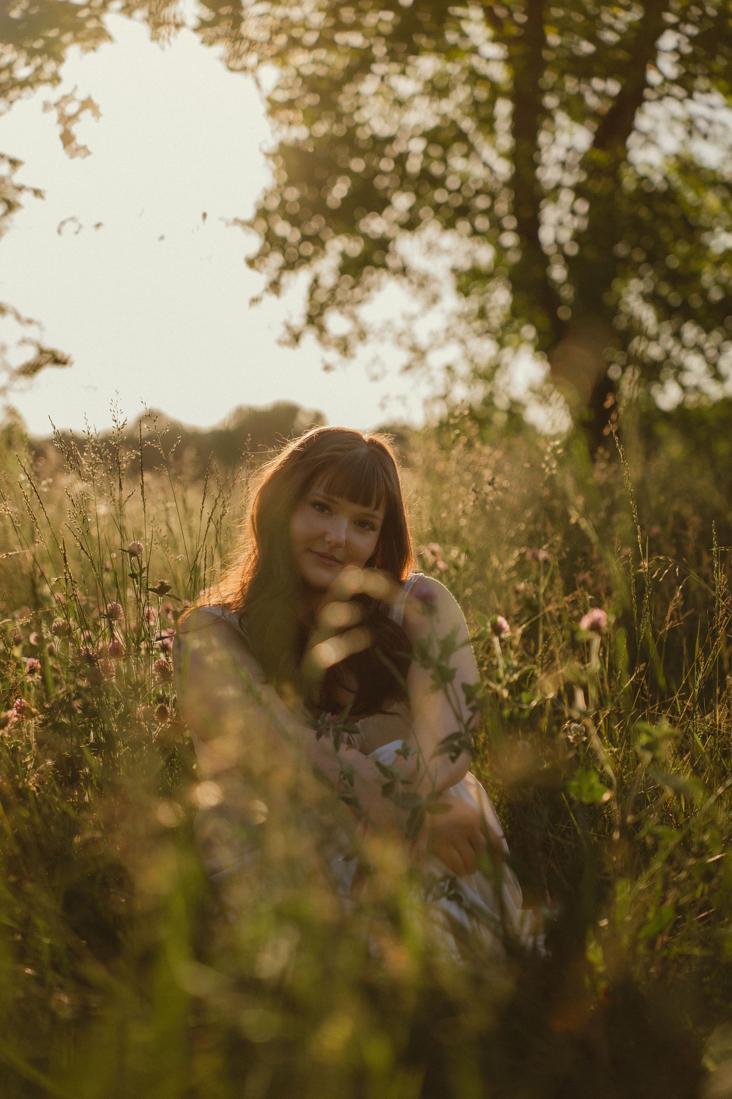 Young woman sitting in a sunlit meadow surrounded by wildflowers and tall grass, smiling softly in golden hour light.