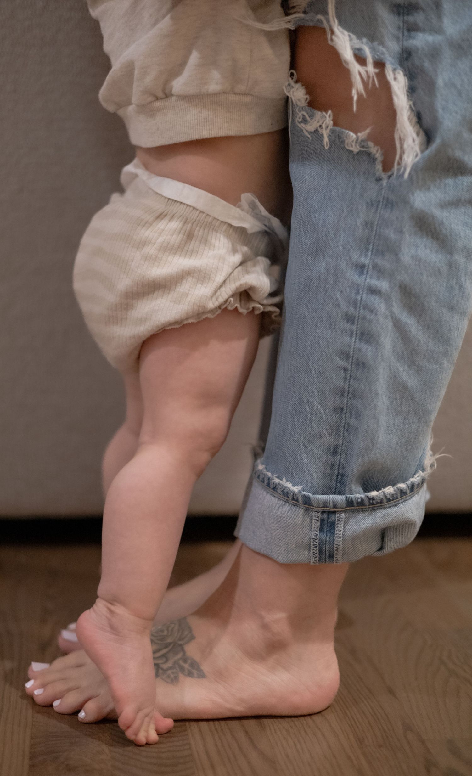 Close-up of baby and barefoot tattooed mother standing together on hardwood floor.