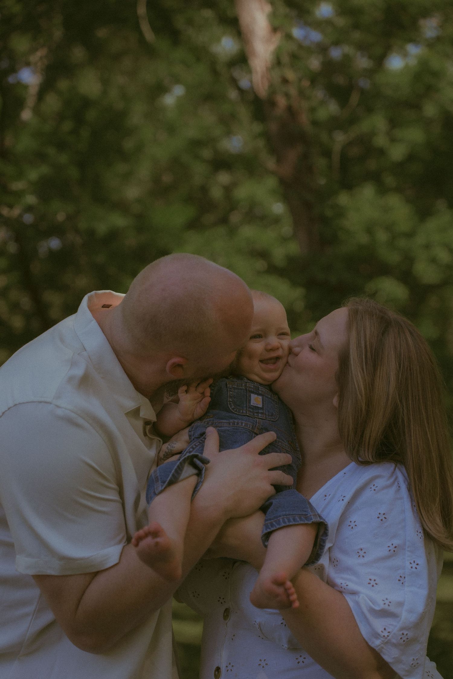 A bald father and blonde mother kiss their laughing baby in denim overalls outdoors among green trees.