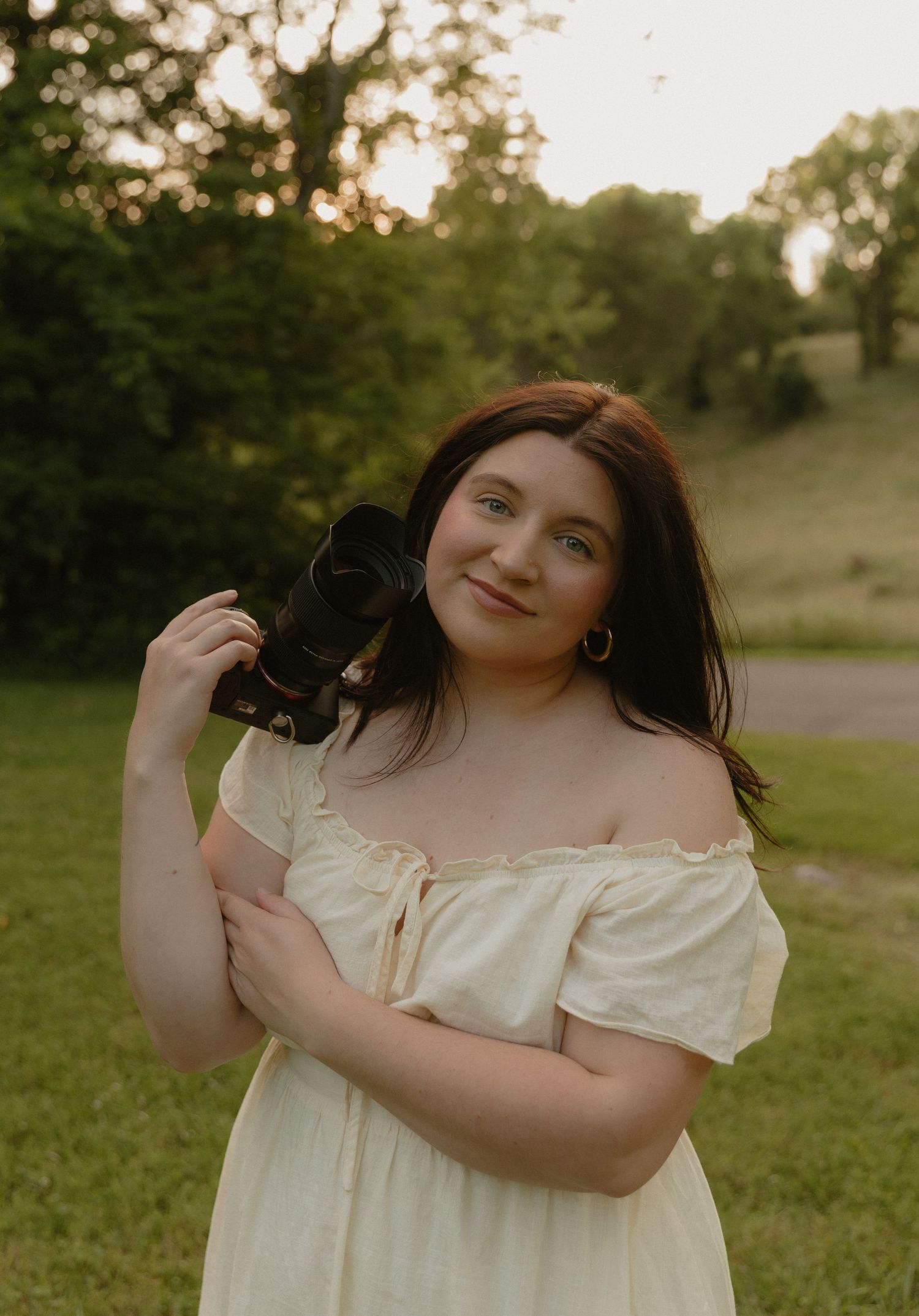 Young woman in off-shoulder cream dress holding a camera outdoors in a lush green park at golden hour.