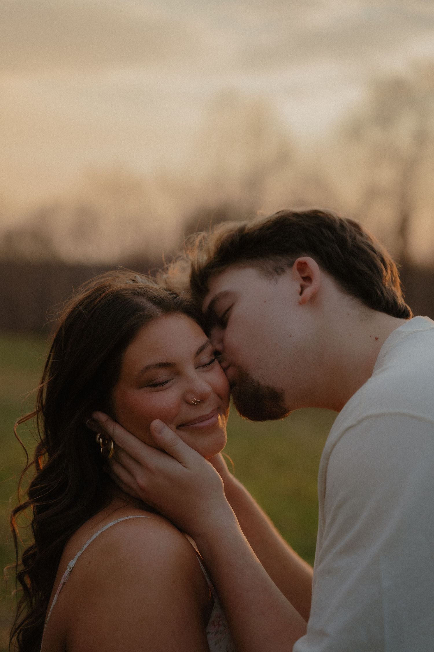 A couple shares a tender sunset kiss outdoors, the man gently cupping the smiling woman's face in a romantic golden hour moment.