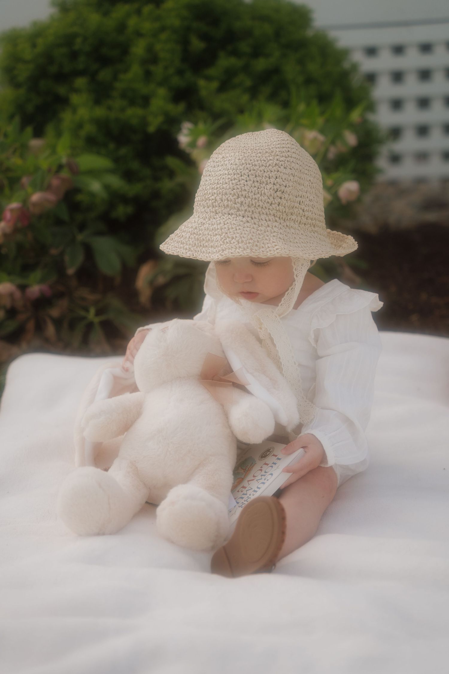 A baby in a straw hat holds a white stuffed bunny on a white blanket outdoors.