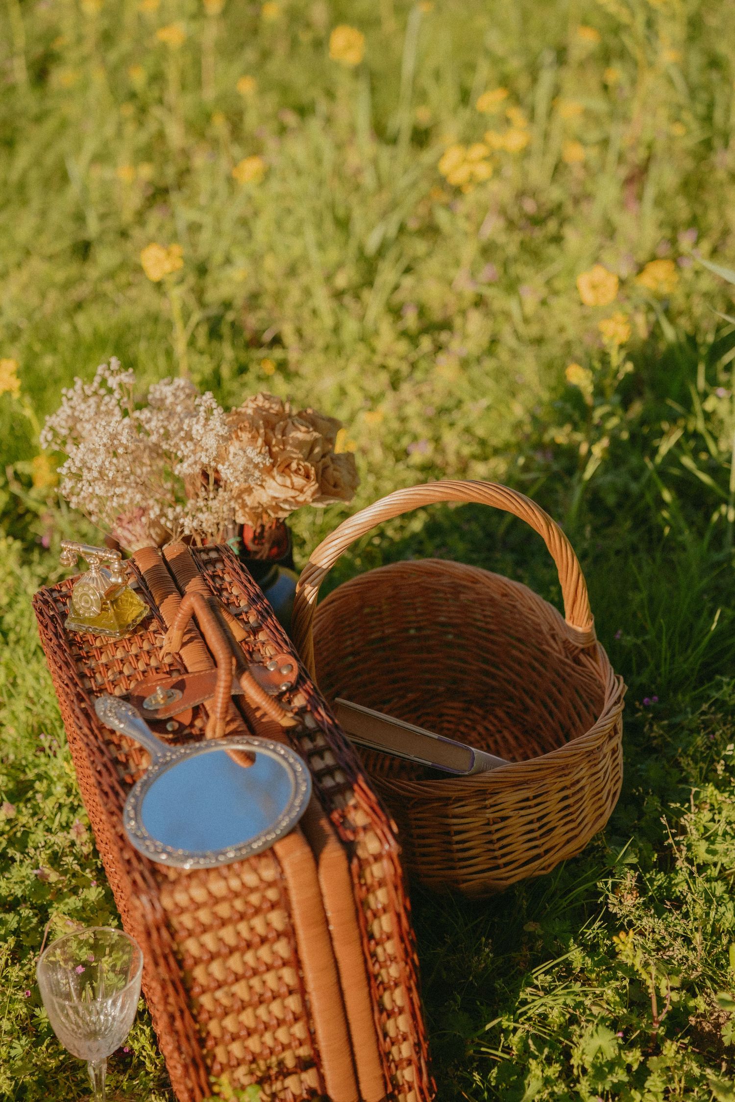 Rustic picnic setup with wicker basket, baguettes, wildflowers, and glassware on a blanket in a sunny meadow.