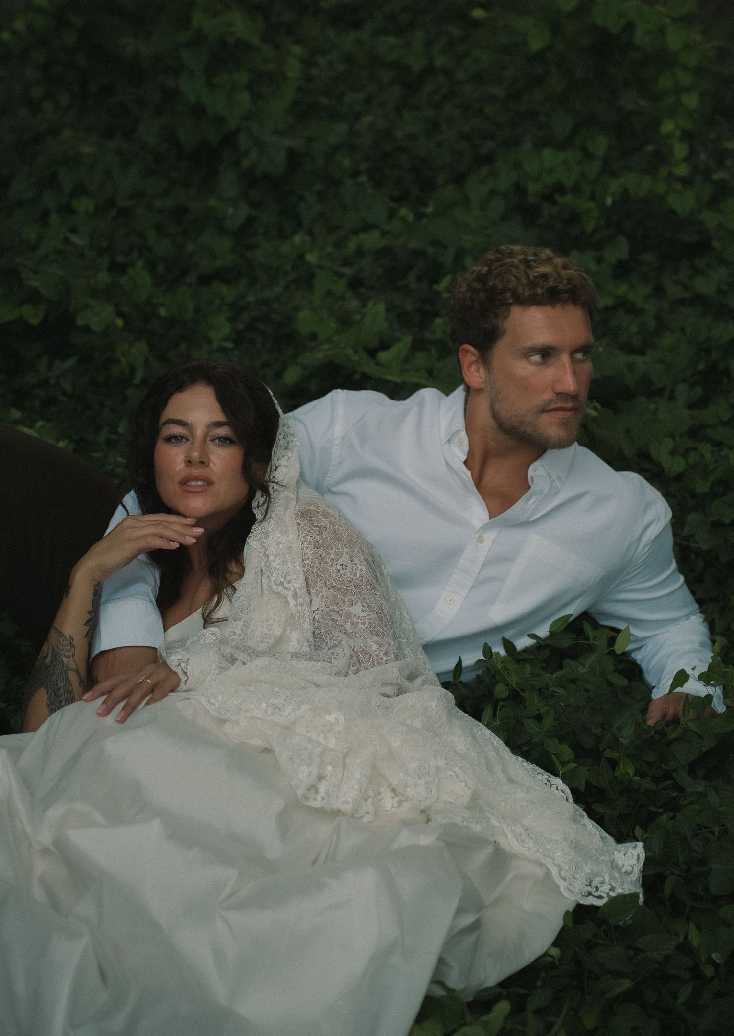 Bride in lace wedding gown sits with groom in white shirt among lush green foliage in romantic outdoor setting.