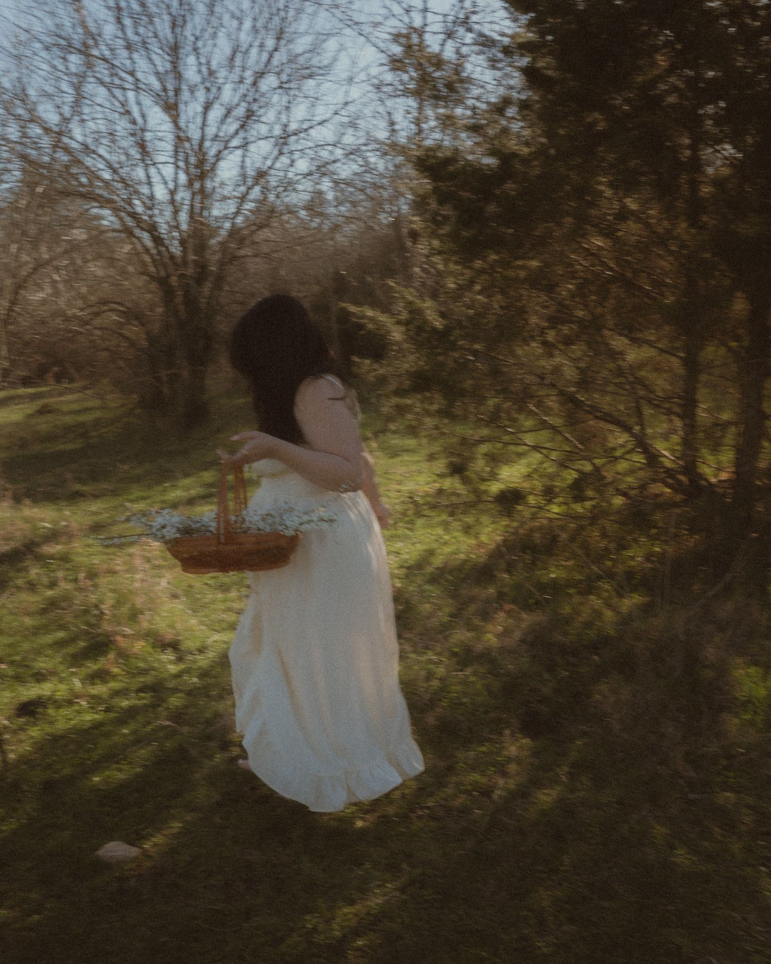 Woman in white dress carrying a wicker flower basket walking through a sunlit green field.
