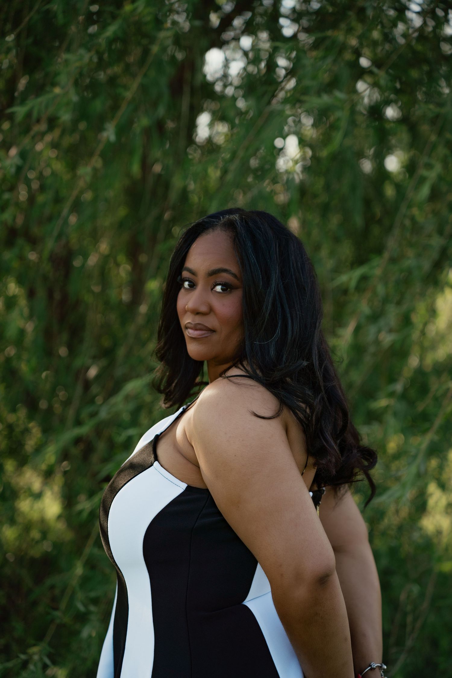 A woman in a black and white sleeveless dress poses outdoors with lush green trees in the background.