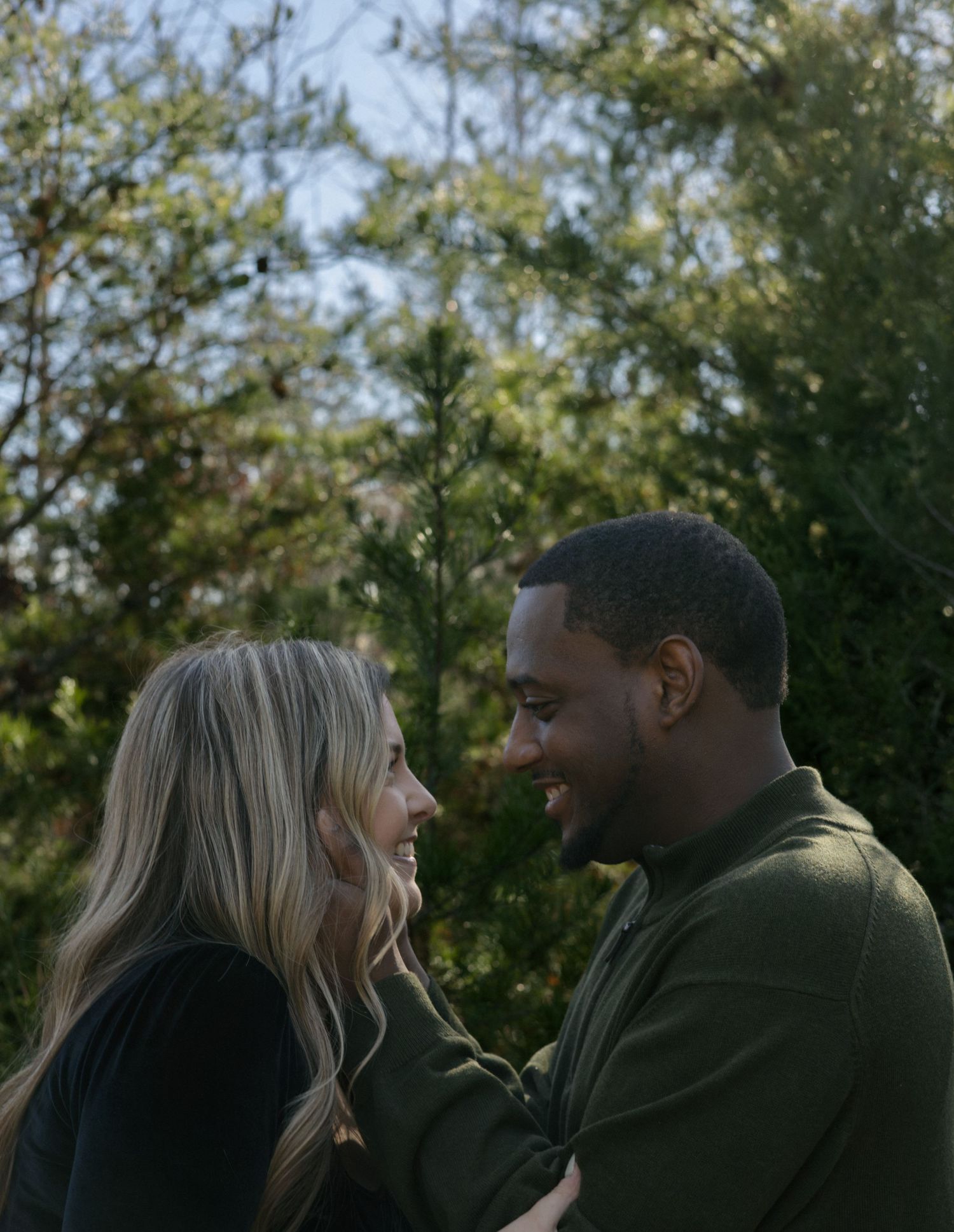 Happy interracial couple smiling at each other closely outdoors surrounded by lush green trees.