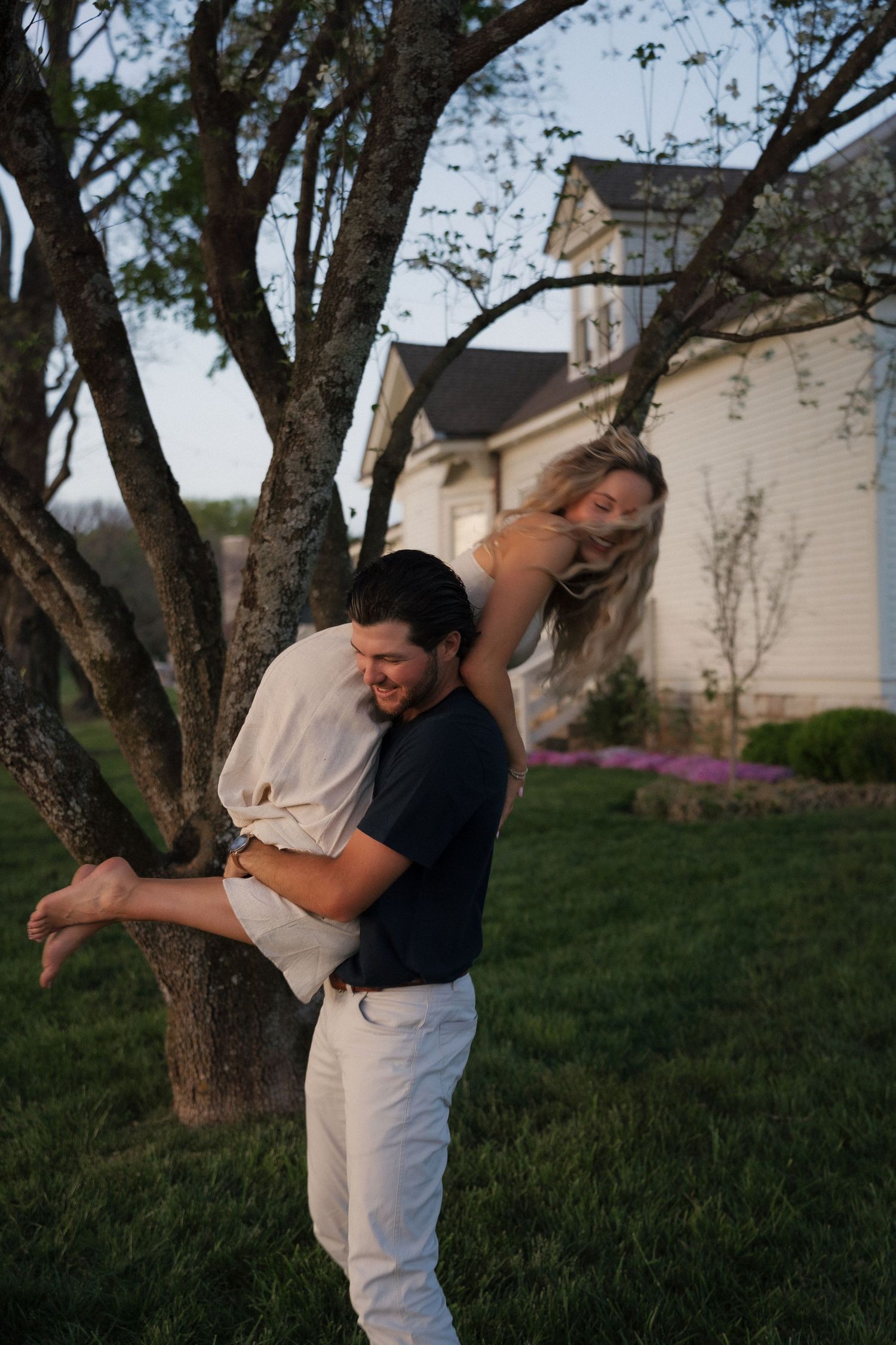 Man playfully lifting laughing blonde woman beside tree in front of white farmhouse at dusk.