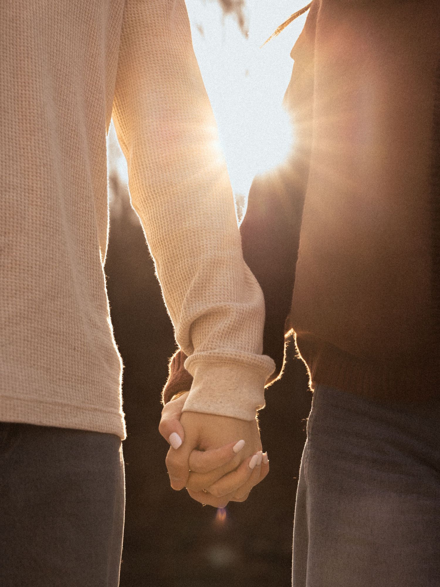Couple holding hands at sunset with warm golden backlight creating a romantic silhouette effect.