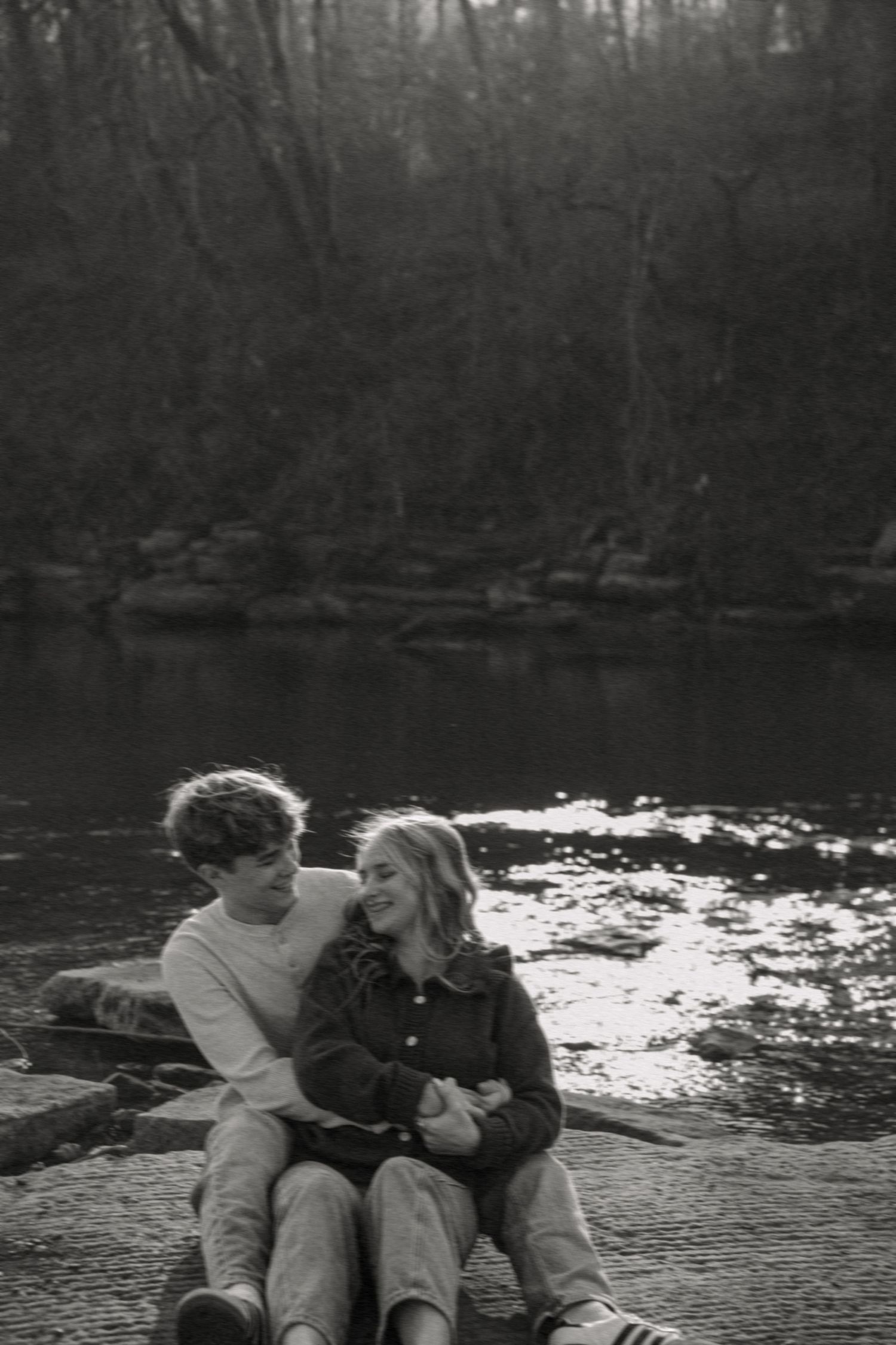 Black and white photo of a couple sitting close together by a reflective river surrounded by bare winter trees.