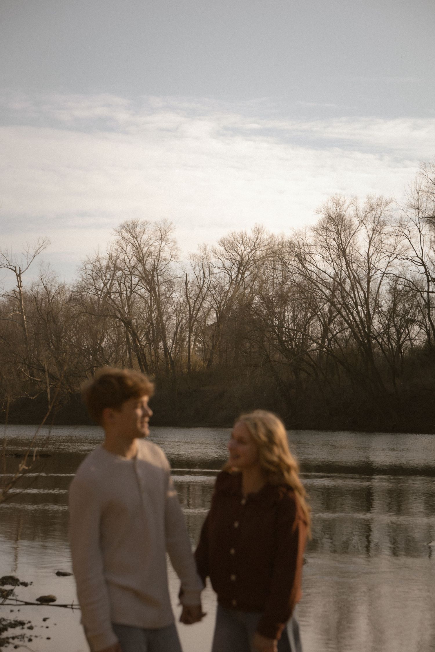 Young couple holding hands near a calm reflective river with bare trees under a cloudy sky.