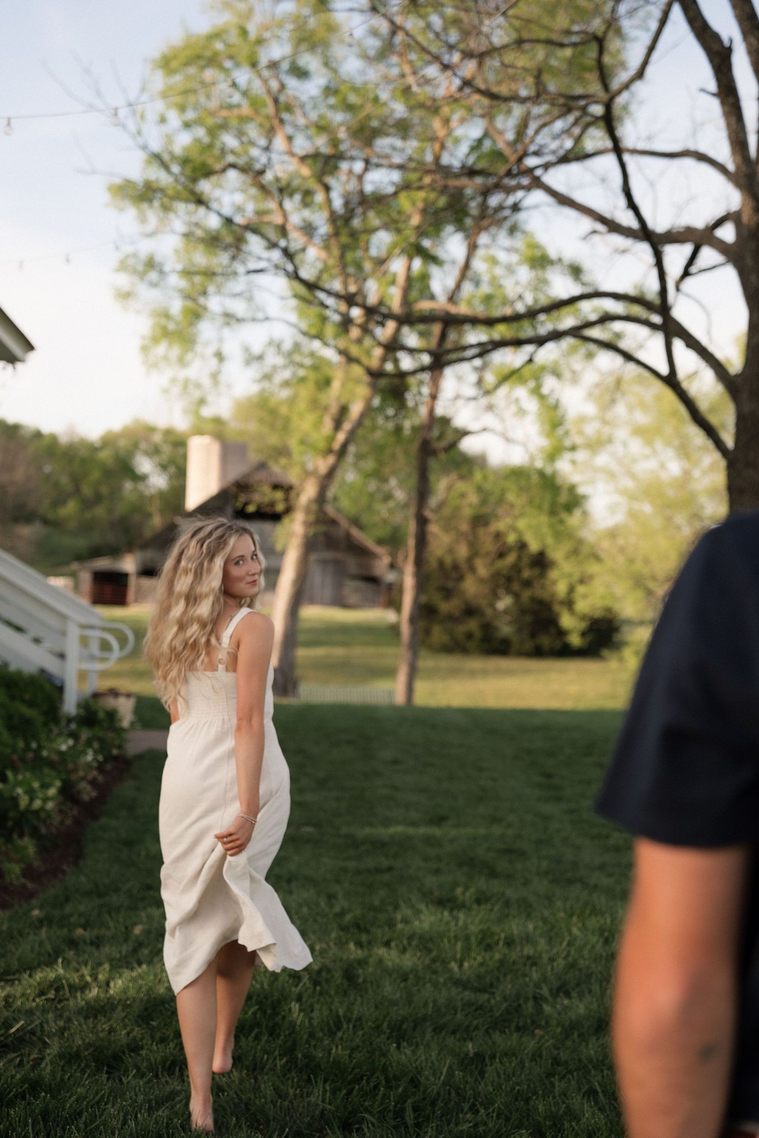 Barefoot blonde woman in white dress twirling on green lawn at golden hour near farmhouse.
