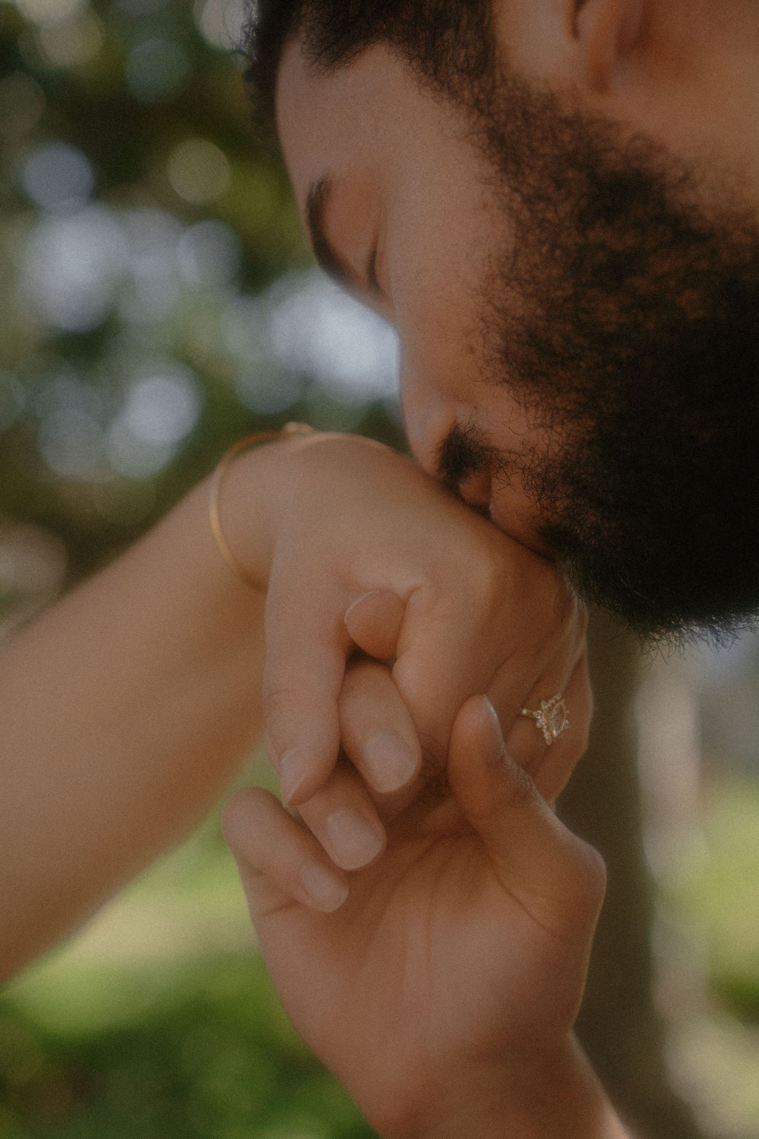 Bearded man gently kissing a woman's hand adorned with a gold ring and bracelet outdoors.