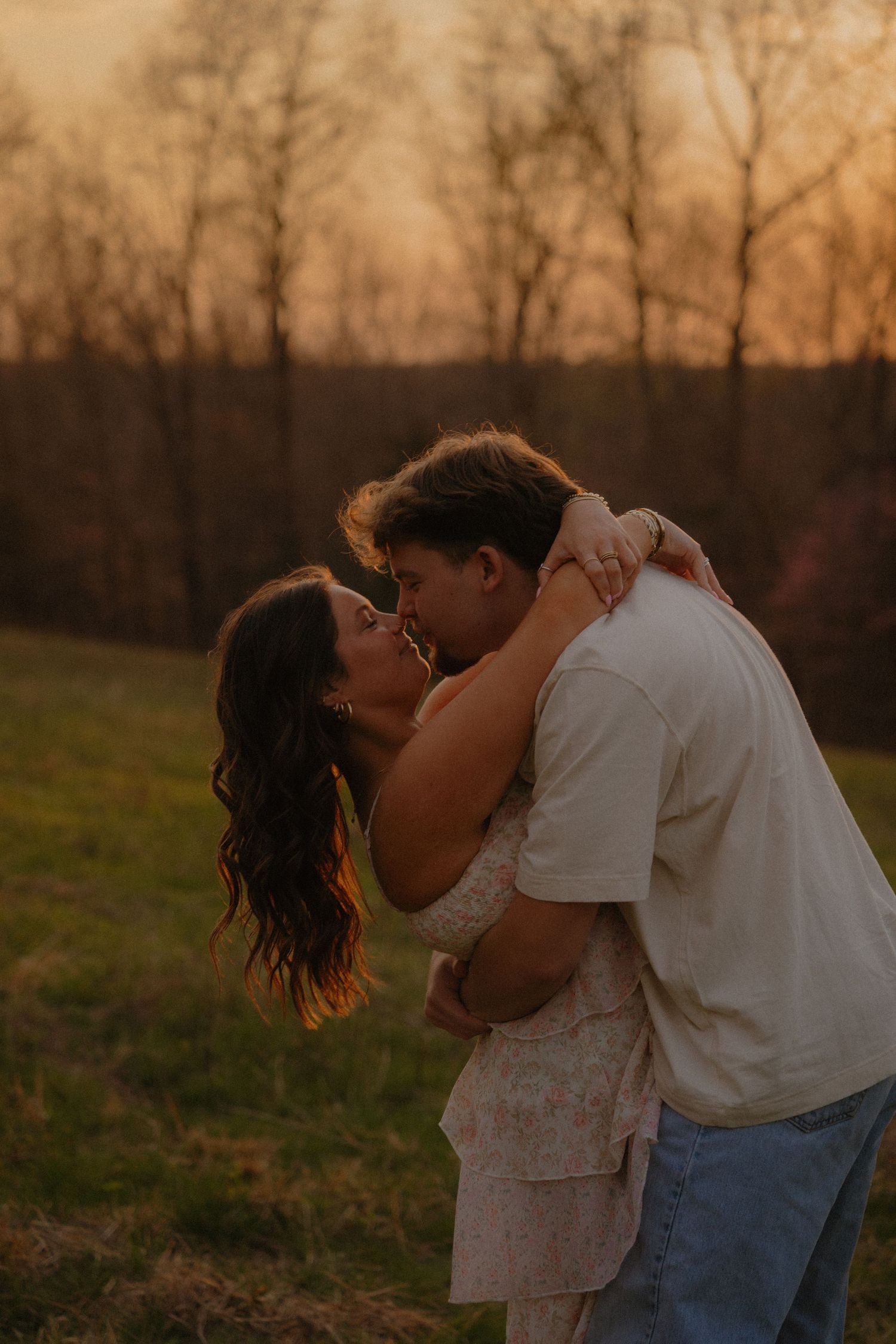 Couple kissing at sunset in an open field with warm orange sky and bare trees in background.