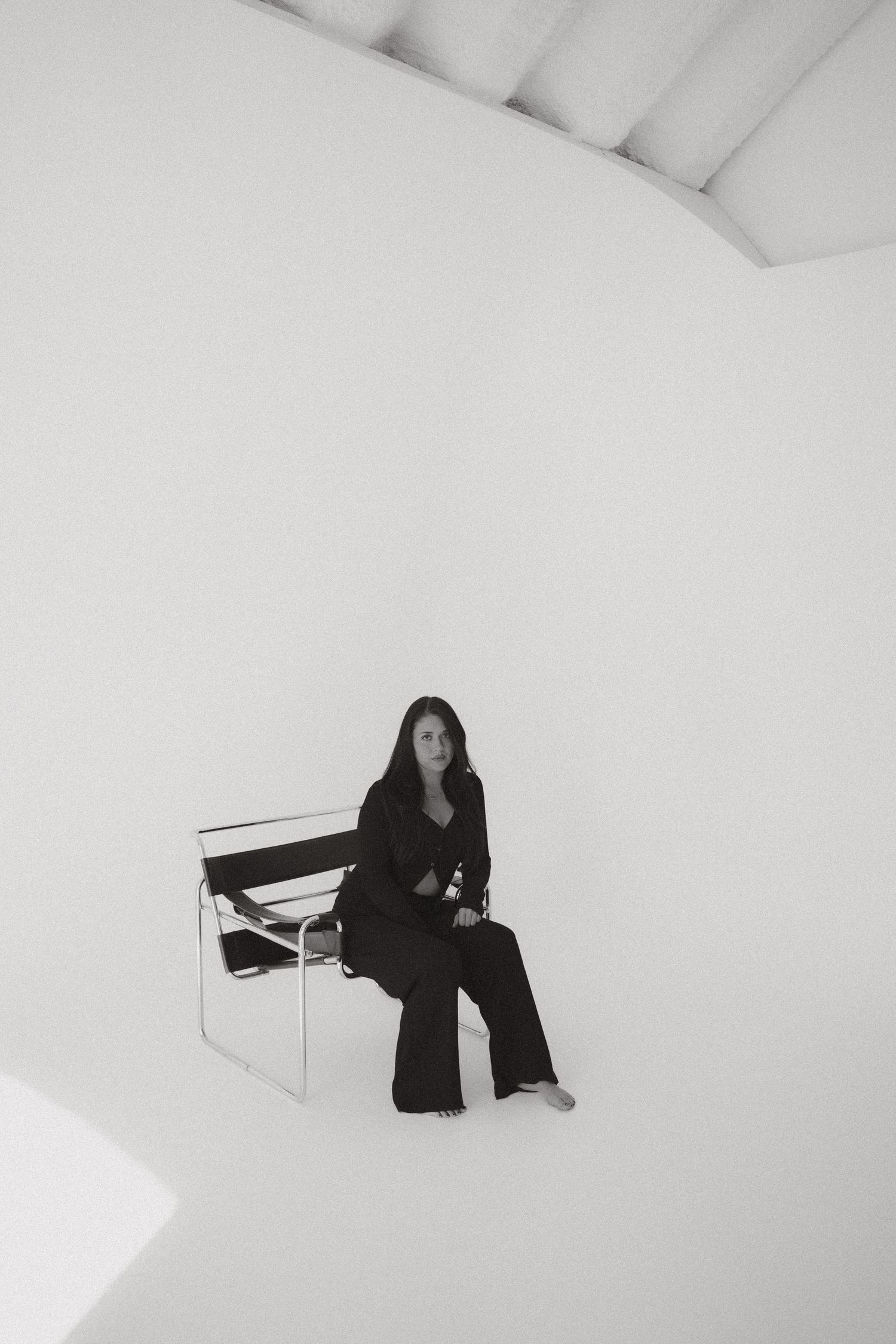 Woman in black outfit sitting on a Wassily chair in a bright white minimalist studio space.
