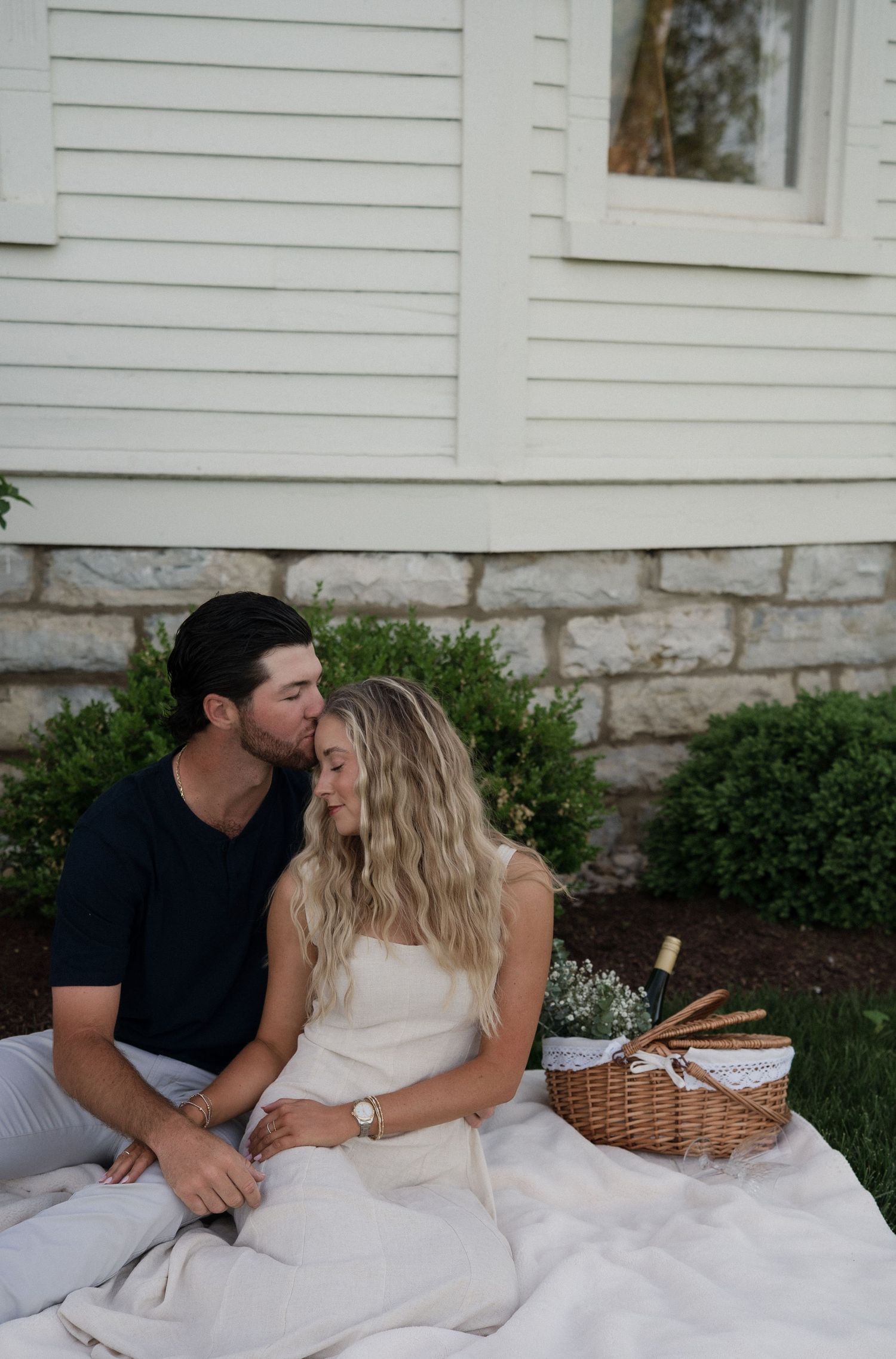 Couple sharing a tender kiss during a romantic outdoor picnic on a white blanket beside a charming house.