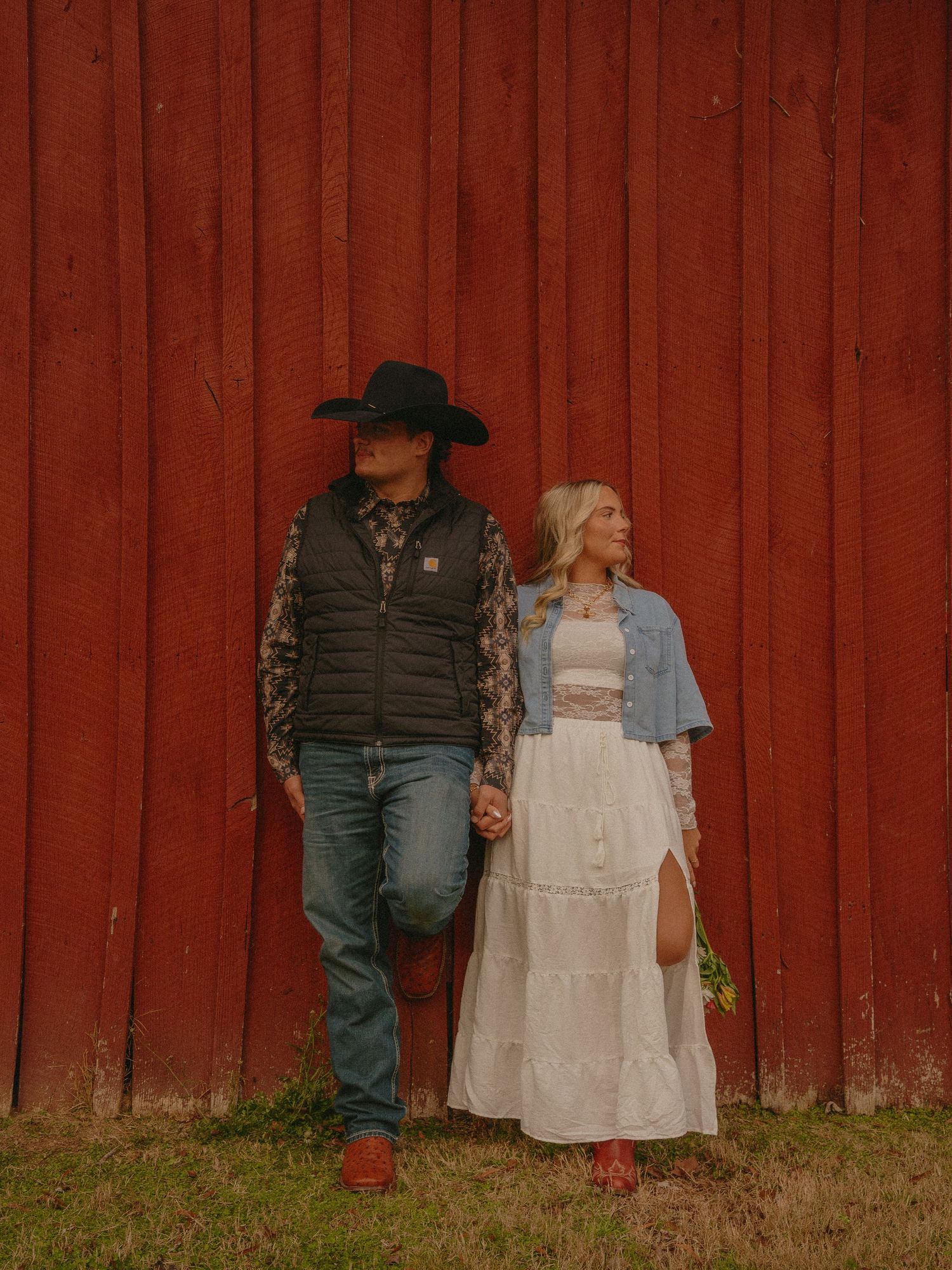 Couple holding hands in front of a red barn wall in western-style engagement photo.