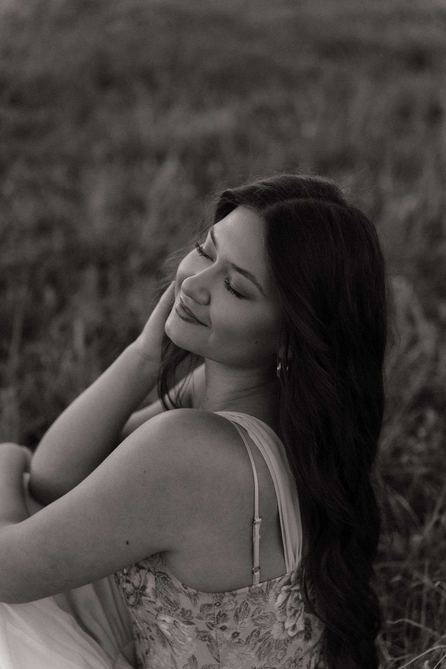 Black and white portrait of dark-haired woman in floral dress sitting in field, eyes closed.
