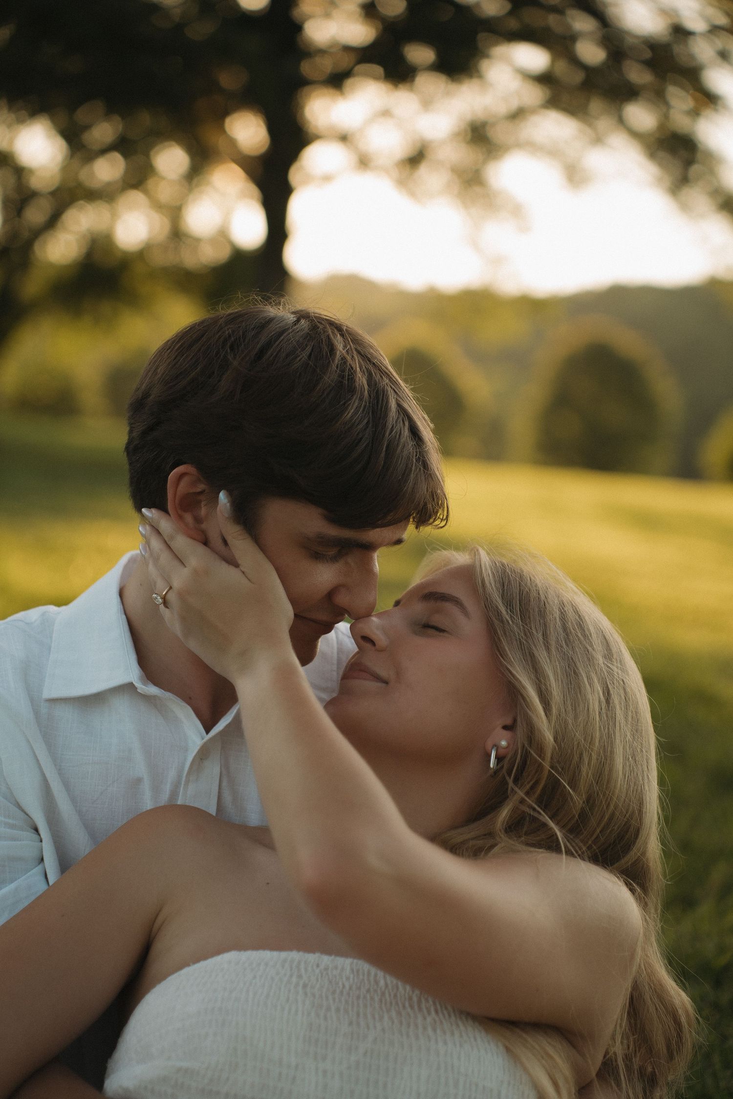 Couple sharing a romantic almost-kiss at golden hour in a lush green field, woman wearing engagement ring.