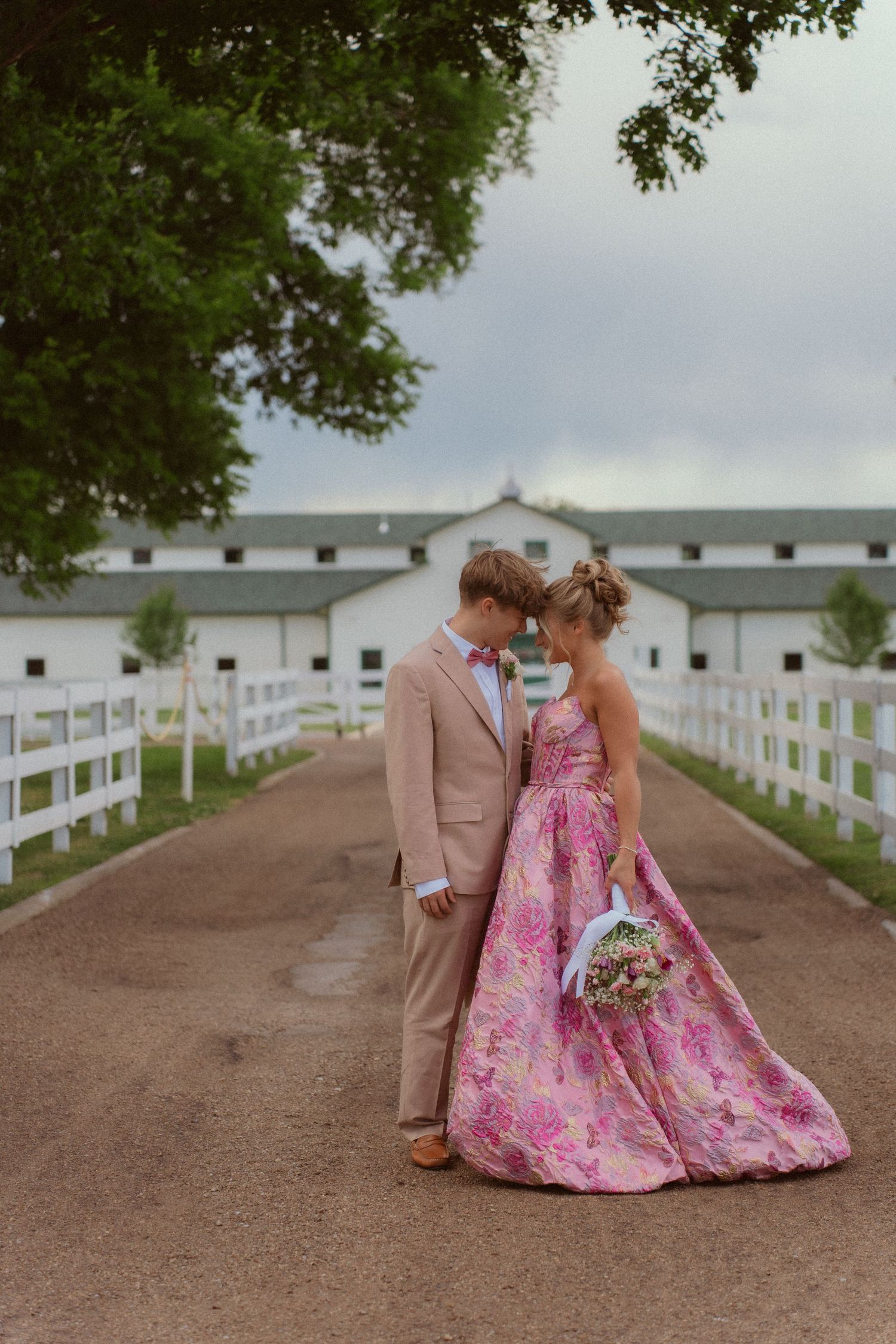 Couple in pink floral gown and tan suit share a romantic moment on a path leading to a white barn.