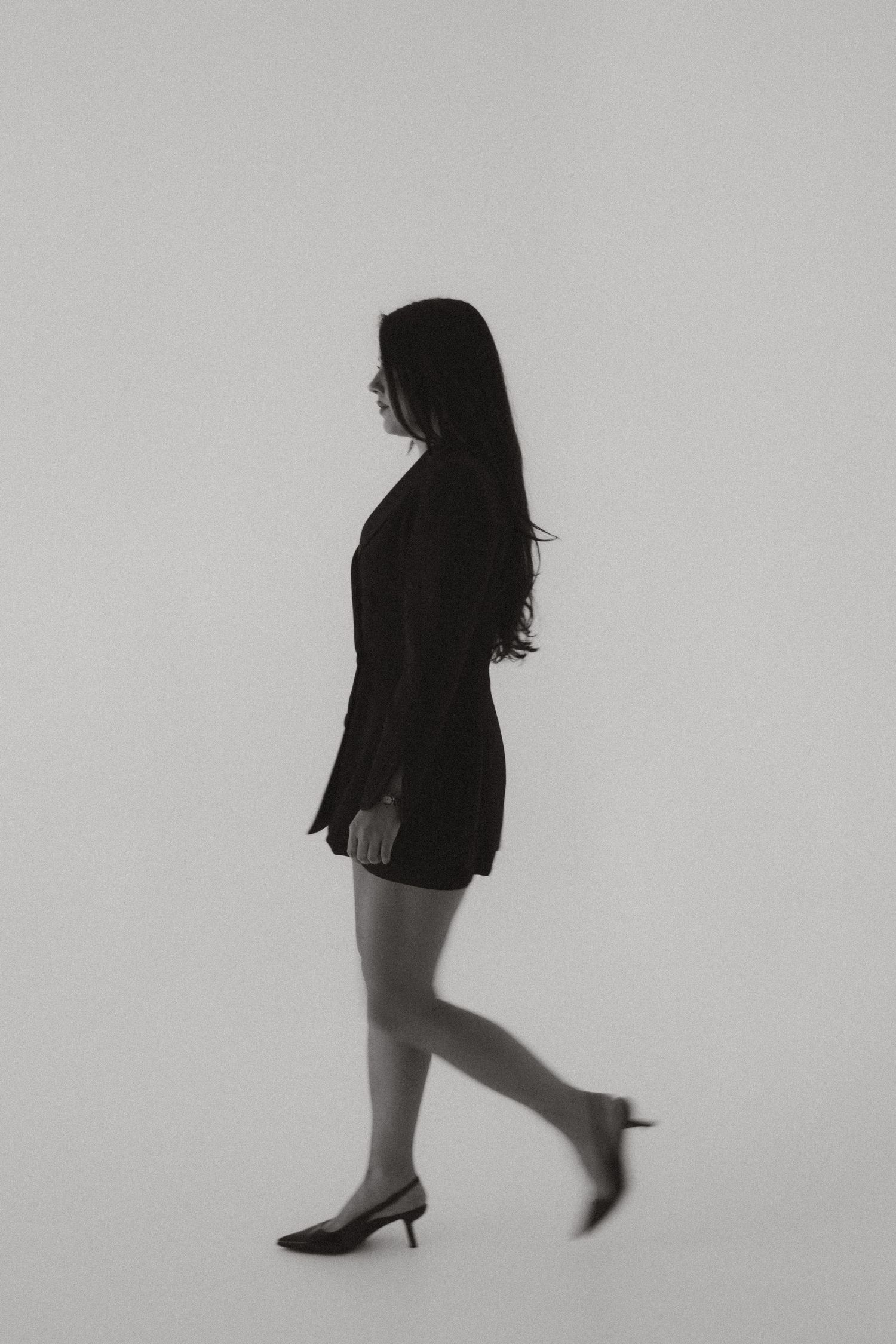 Silhouette of a woman in black mini dress and heels walking against a light background.