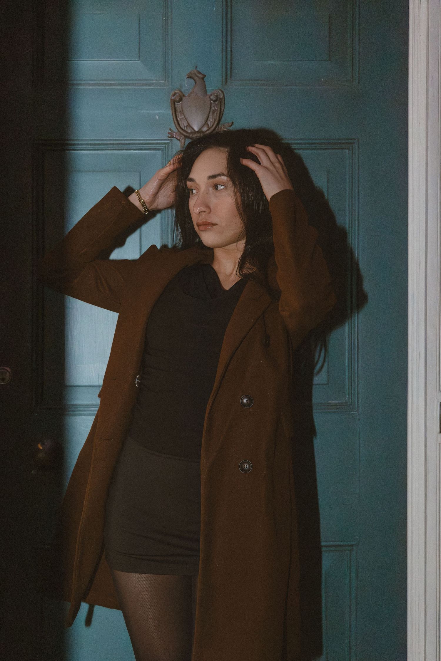Young woman in brown coat posing by a teal door with dramatic sunlight and shadows.